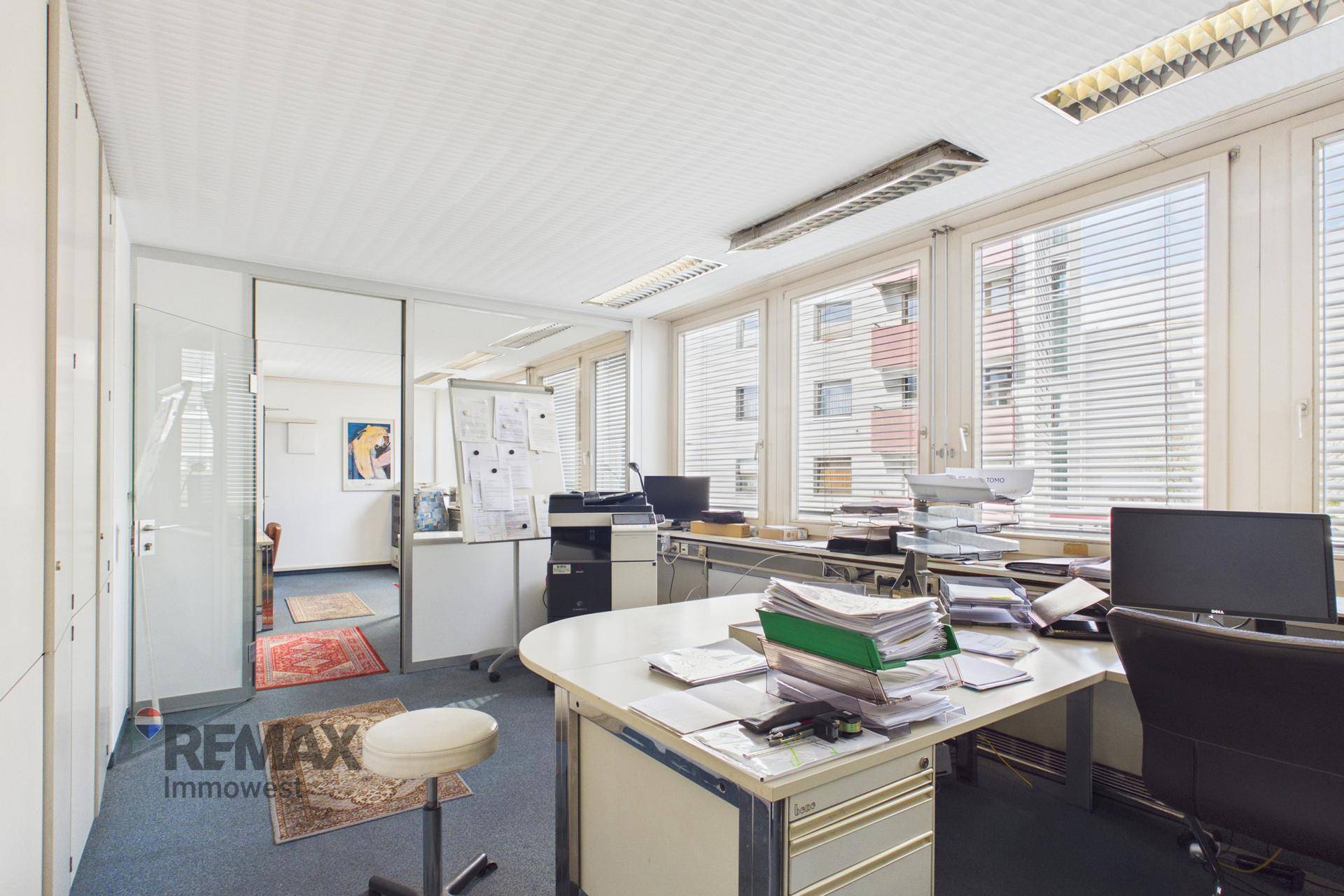 Büroarbeitsplatz mit Schreibtisch, Computer, weißen Schränken und Fenstern mit Jalousien.