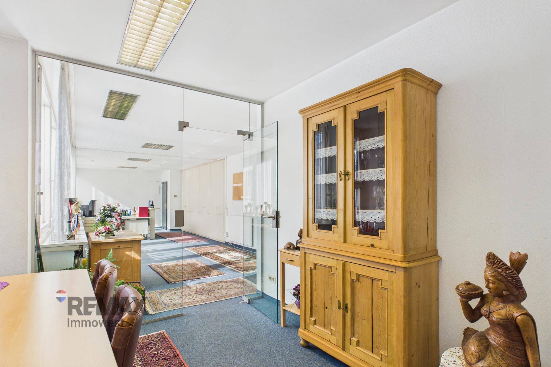 Büroraum mit Glastrennwand, traditionellem Holzschrank und blauem Teppichboden.