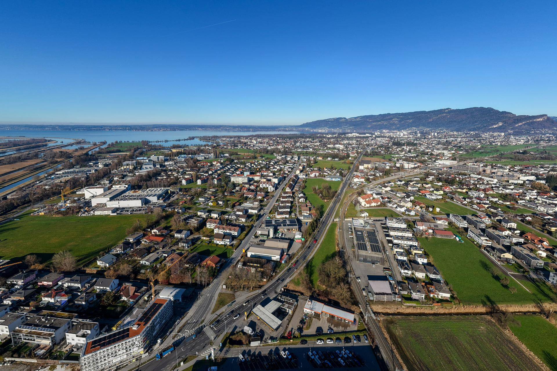 Panoramablick aus der Vogelperspektive auf eine Stadtlandschaft mit See, Bergen und weitläufigen Grünflächen.