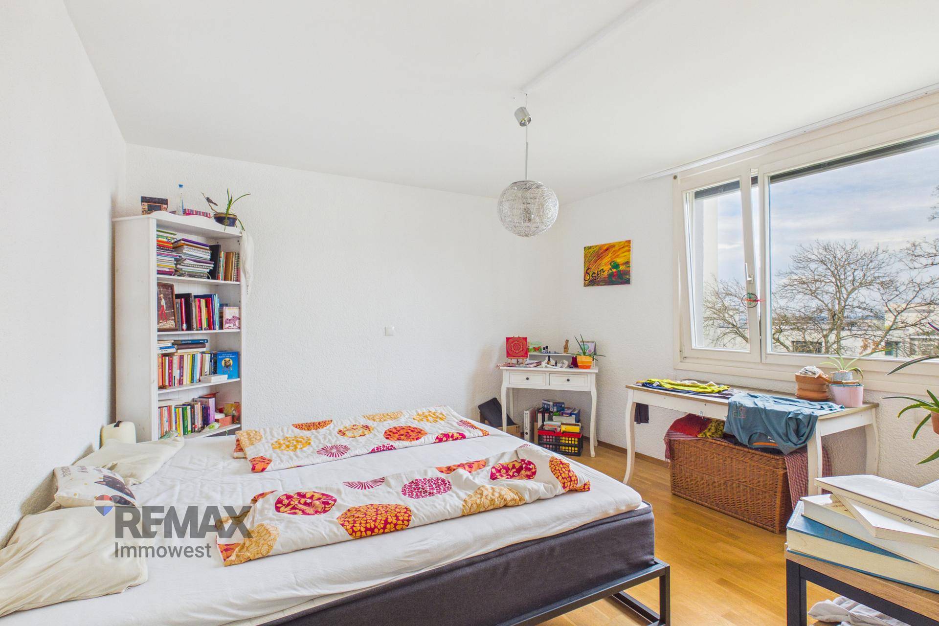 Helles Schlafzimmer mit Parkettboden, großem Fenster und einfacher Einrichtung.