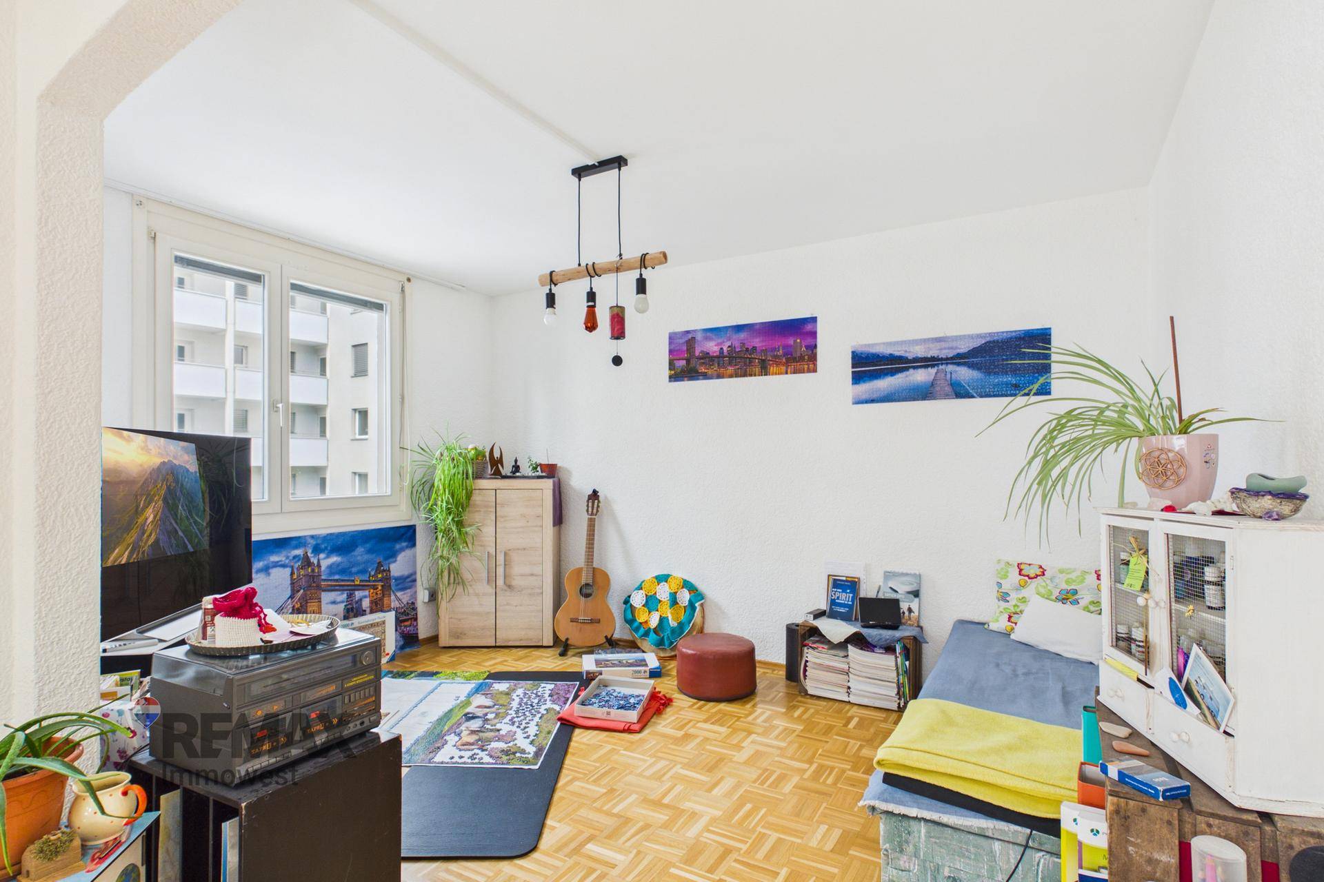 Wohnzimmer mit Parkettboden, Fernseher und Blick auf die umliegenden Gebäude.