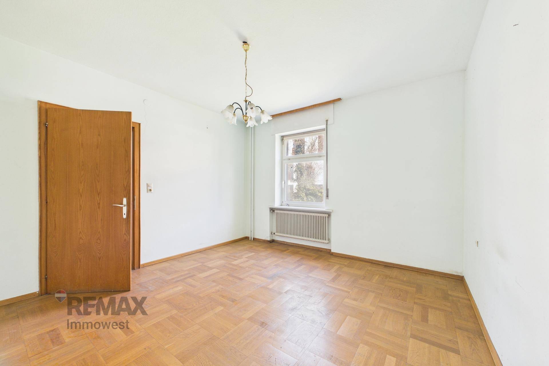 Leeres Zimmer mit Fischgrätparkettboden und einem Fenster mit Heizkörper darunter.