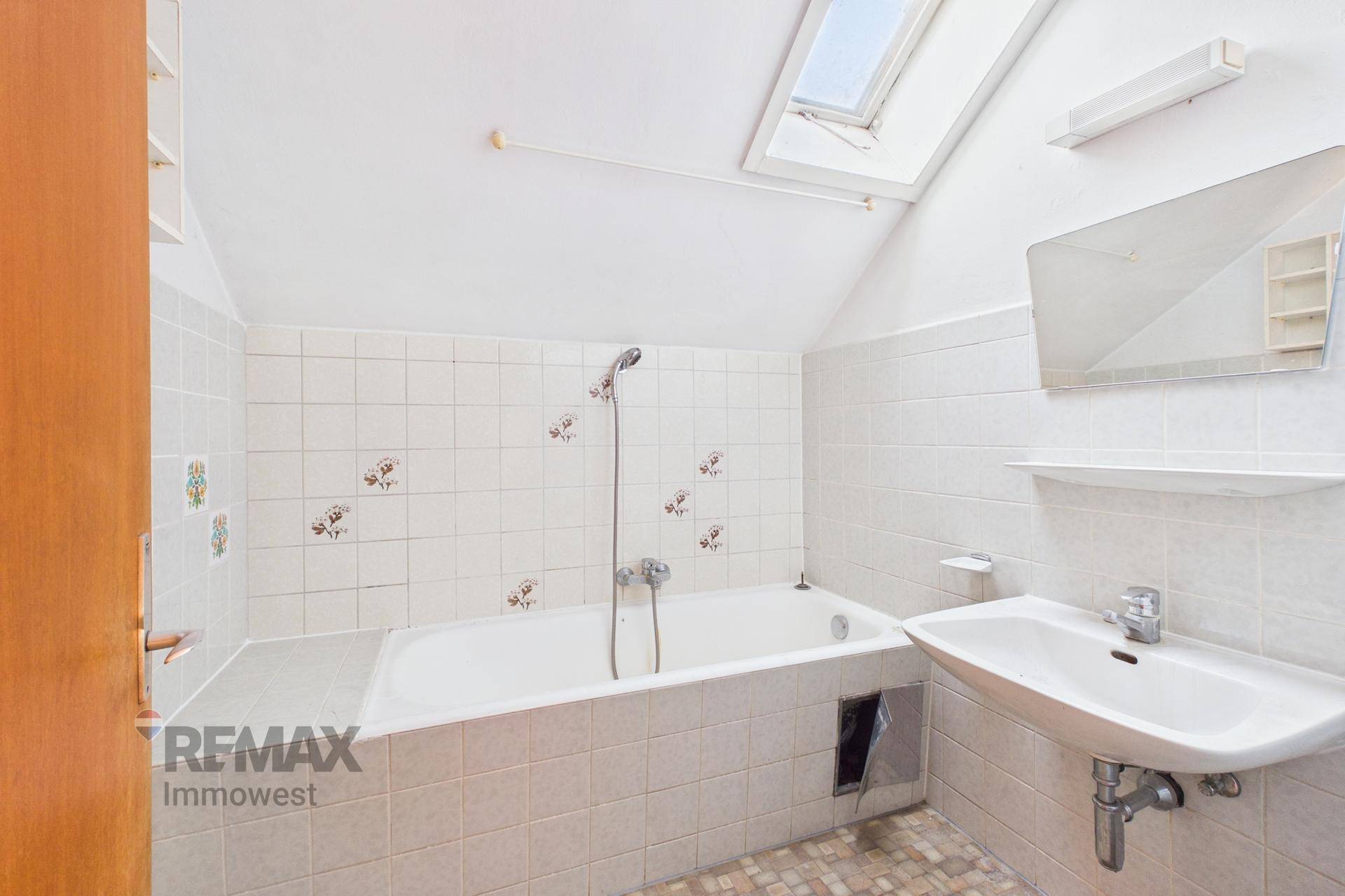 Badezimmer mit Badewanne, Waschbecken, weißen Fliesen und einem Dachfenster.