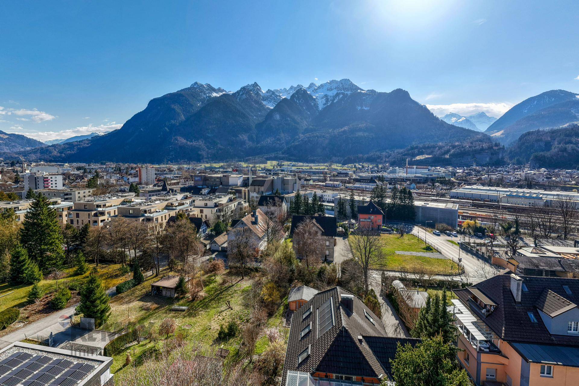 Panoramablick über die Stadt mit Wohngebieten, Grünflächen und der imposanten Bergkette im Hintergrund.