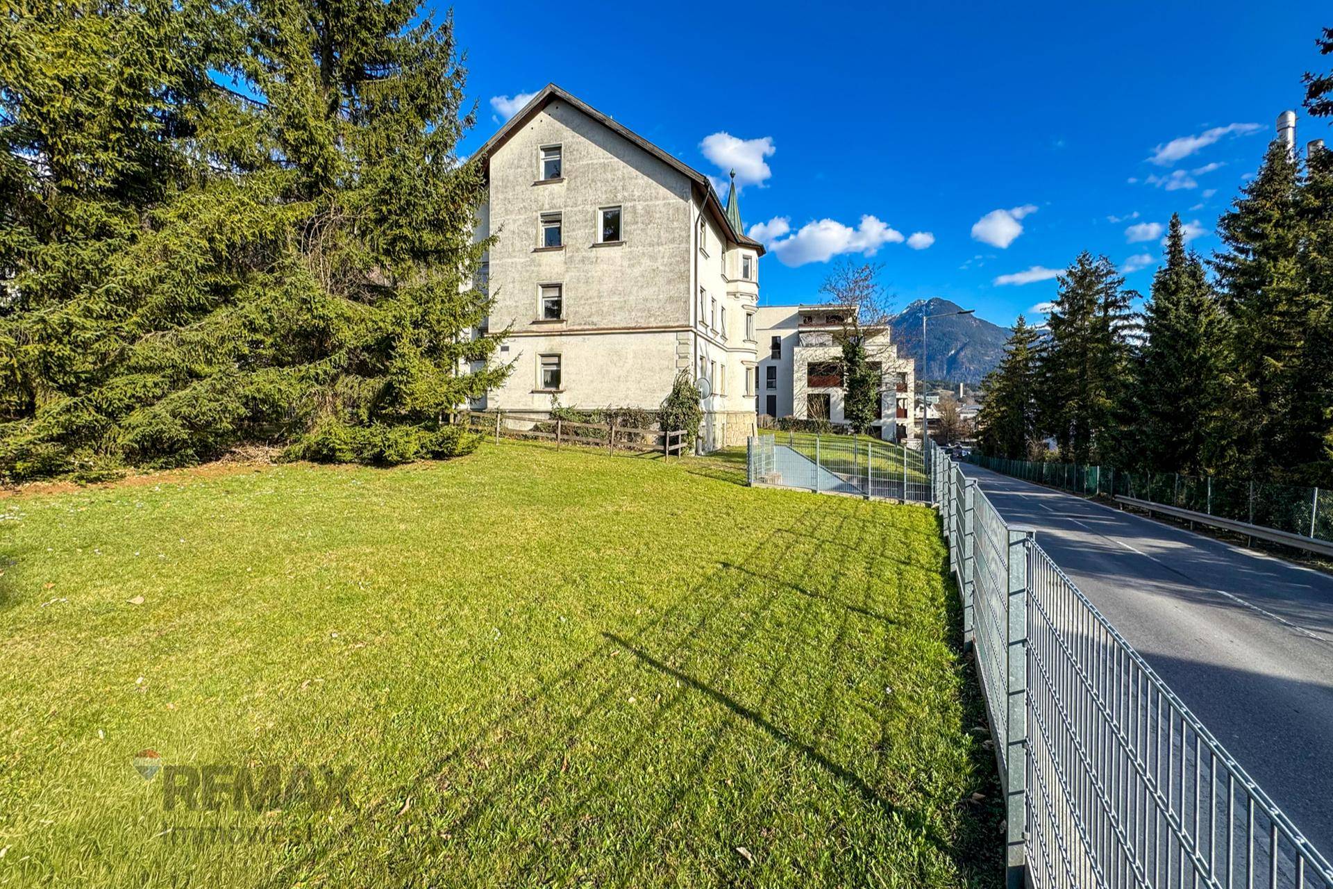 Weitläufiger Garten mit Rasenfläche, Bäumen und Blick auf die Gebäudefassade und die Berge.