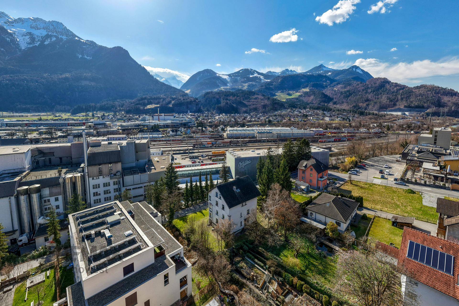Weitläufige Luftaufnahme der Stadt mit Wohngebäuden, Industriegebiet und beeindruckendem Bergpanorama.