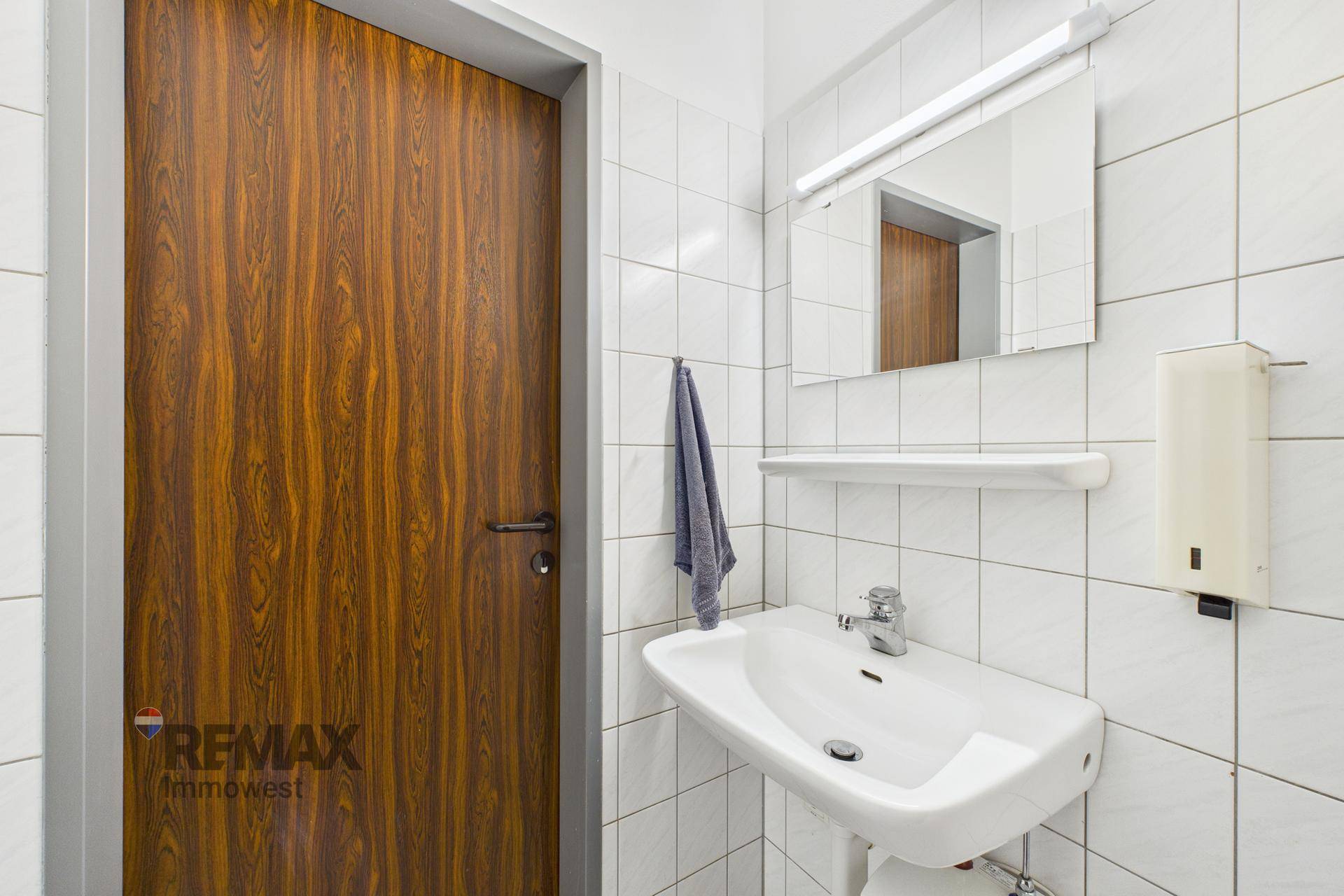 Waschbeckenbereich in einem WC mit Spiegel, Seifenspender und weißen Fliesen.