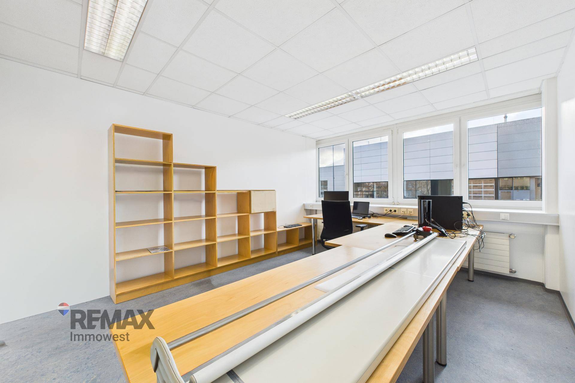 Geräumiges Büro mit langen Schreibtischen und einem Stufenregal an der Wand.