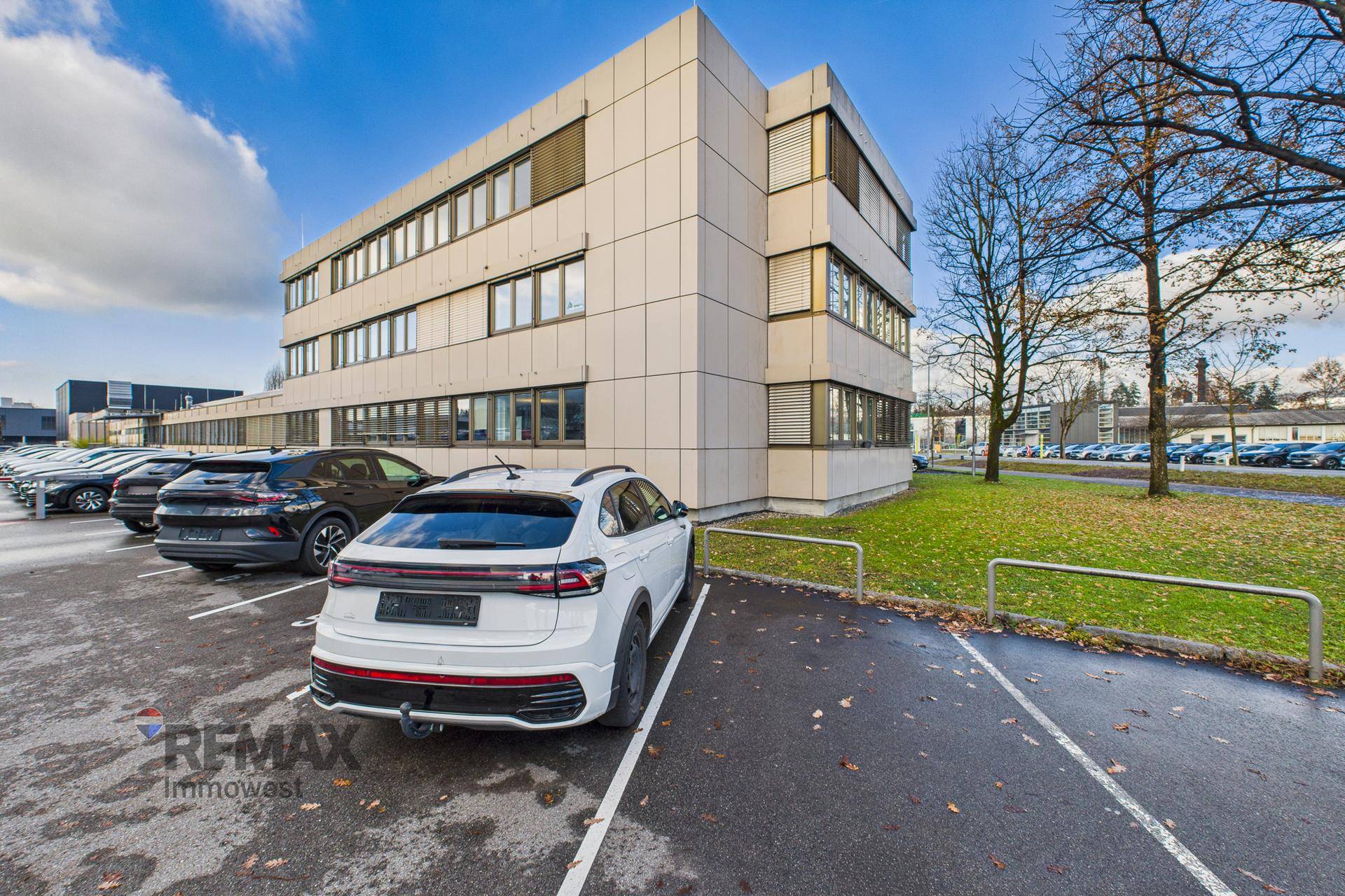 Modernes Bürogebäude mit Parkplätzen und Grünflächen unter blauem Himmel.