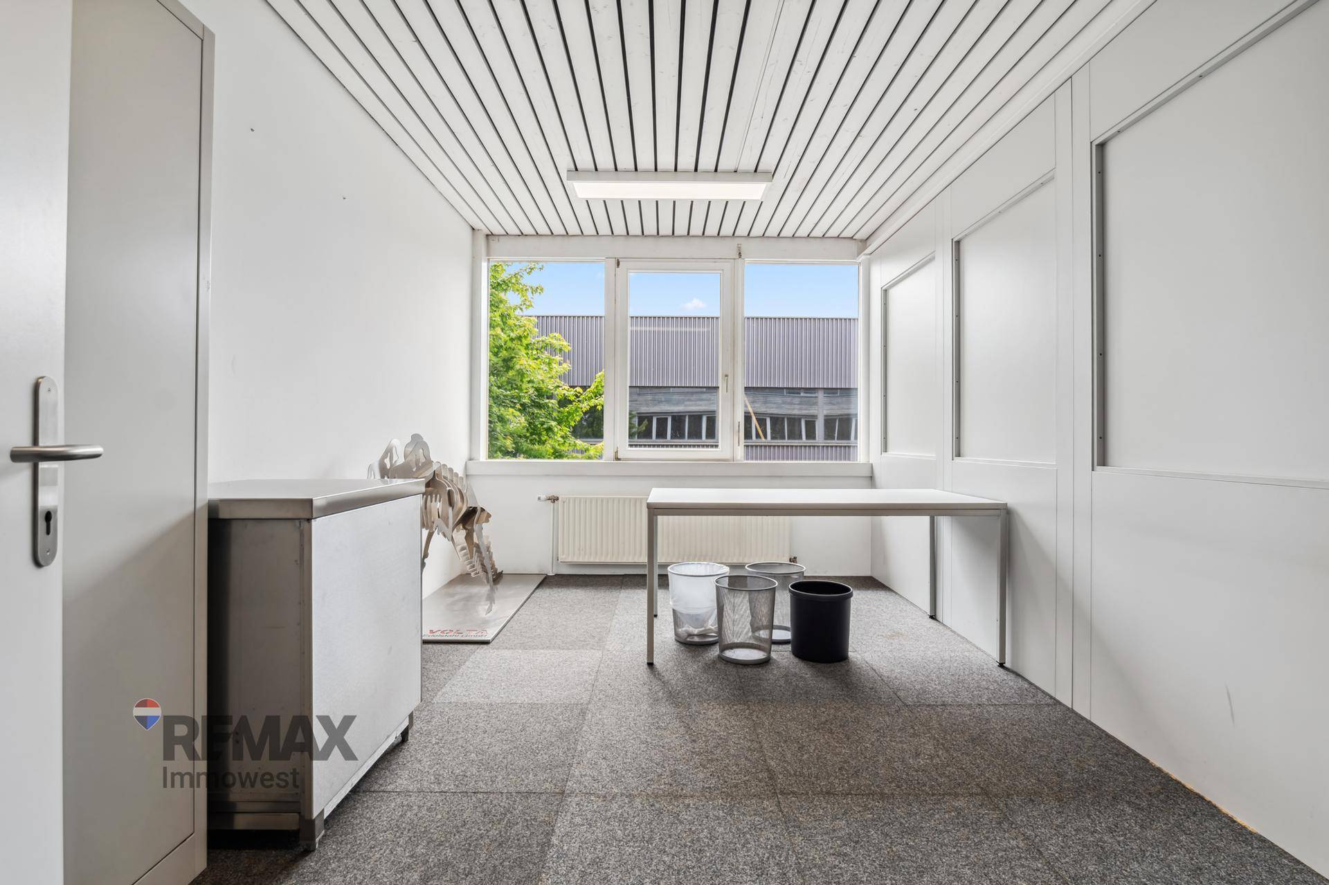 Kleines Büro mit Teppichboden, einem Schreibtisch und großen Fenstern mit Blick ins Grüne.
