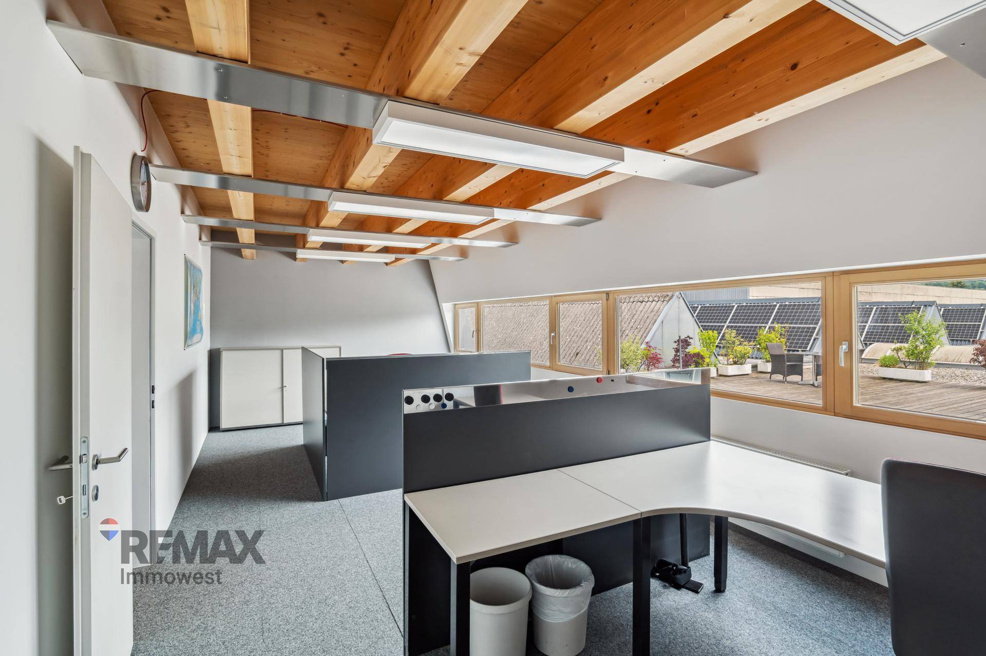 Büro mit Holzbalkendecke, Teppichboden und Blick auf eine Dachterrasse mit Solaranlage.