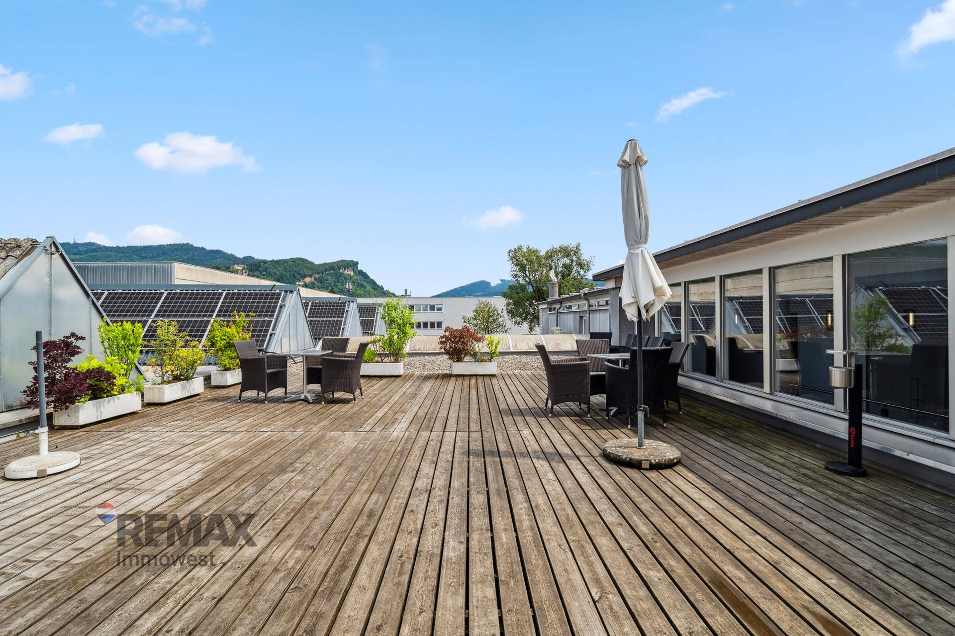 Große Dachterrasse mit Holzboden, Sitzgelegenheiten und Sonnenschirm, umgeben von Pflanzen und Bergen.