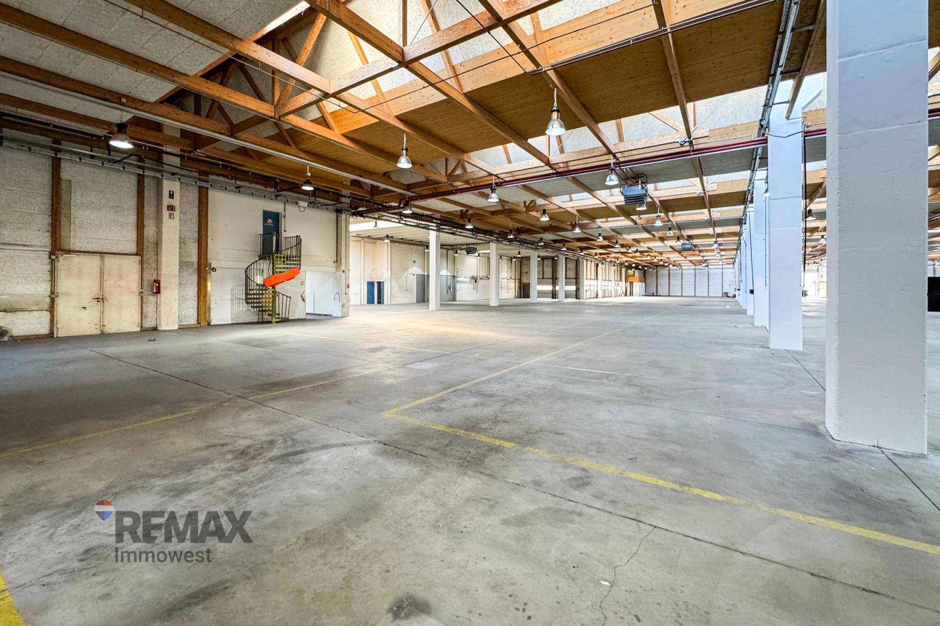 Weitläufige Lagerhalle mit Betonboden, Holzbalkendecke und einer Wendeltreppe im Hintergrund.