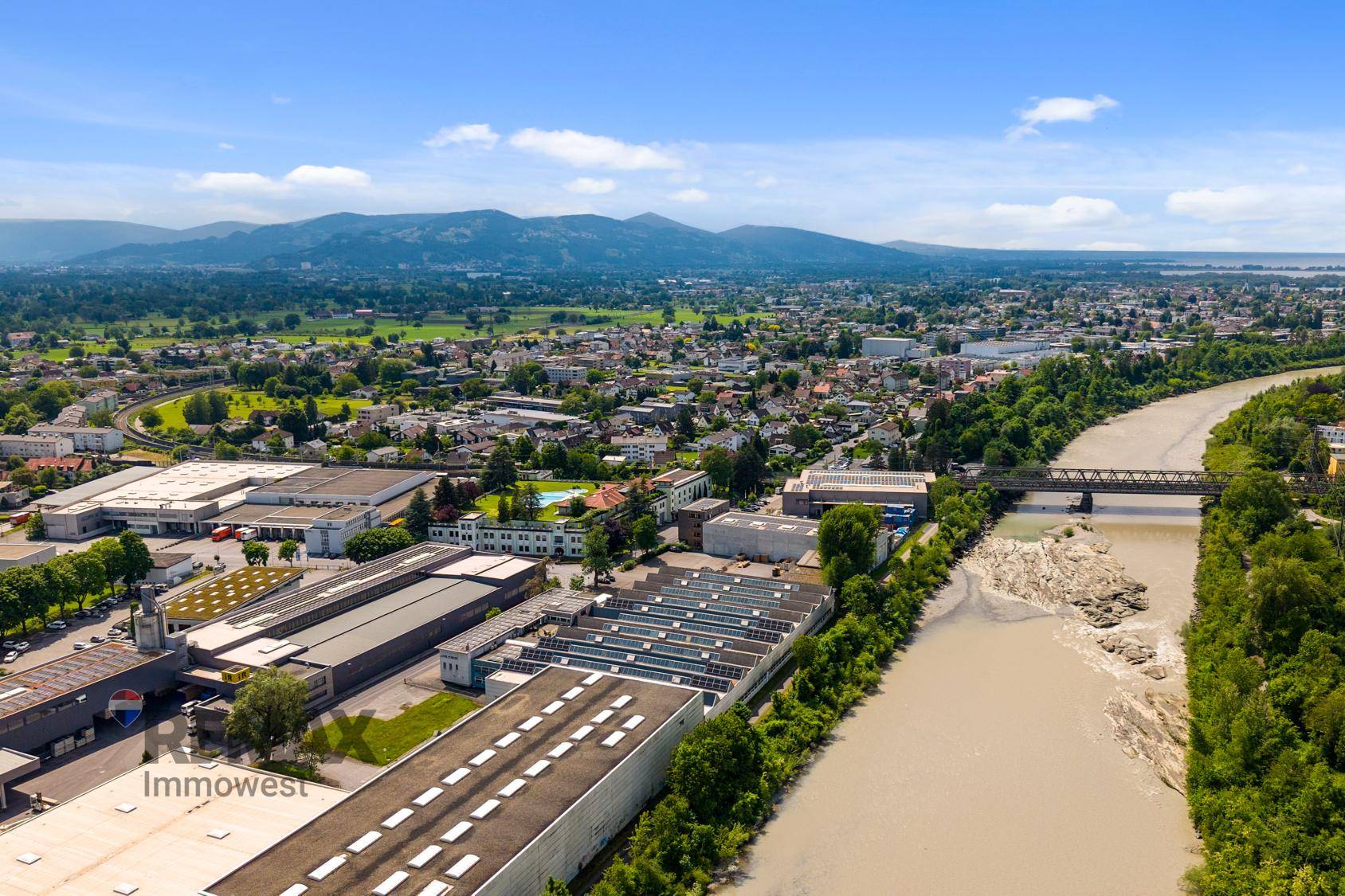 Weitwinkel-Luftaufnahme einer Stadtlandschaft mit Fluss, Industriegebiet und einer beeindruckenden Bergkulisse.