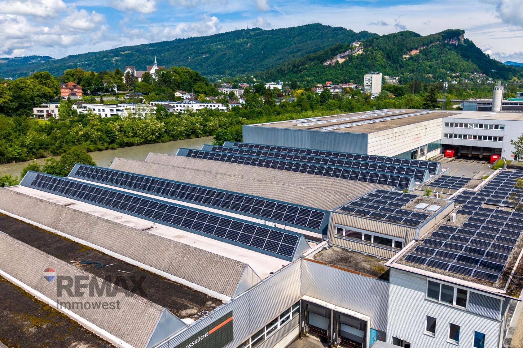 Luftaufnahme eines Industriegebäudes mit zahlreichen Solarmodulen auf den Dächern und grüner Berglandschaft im Hintergrund.