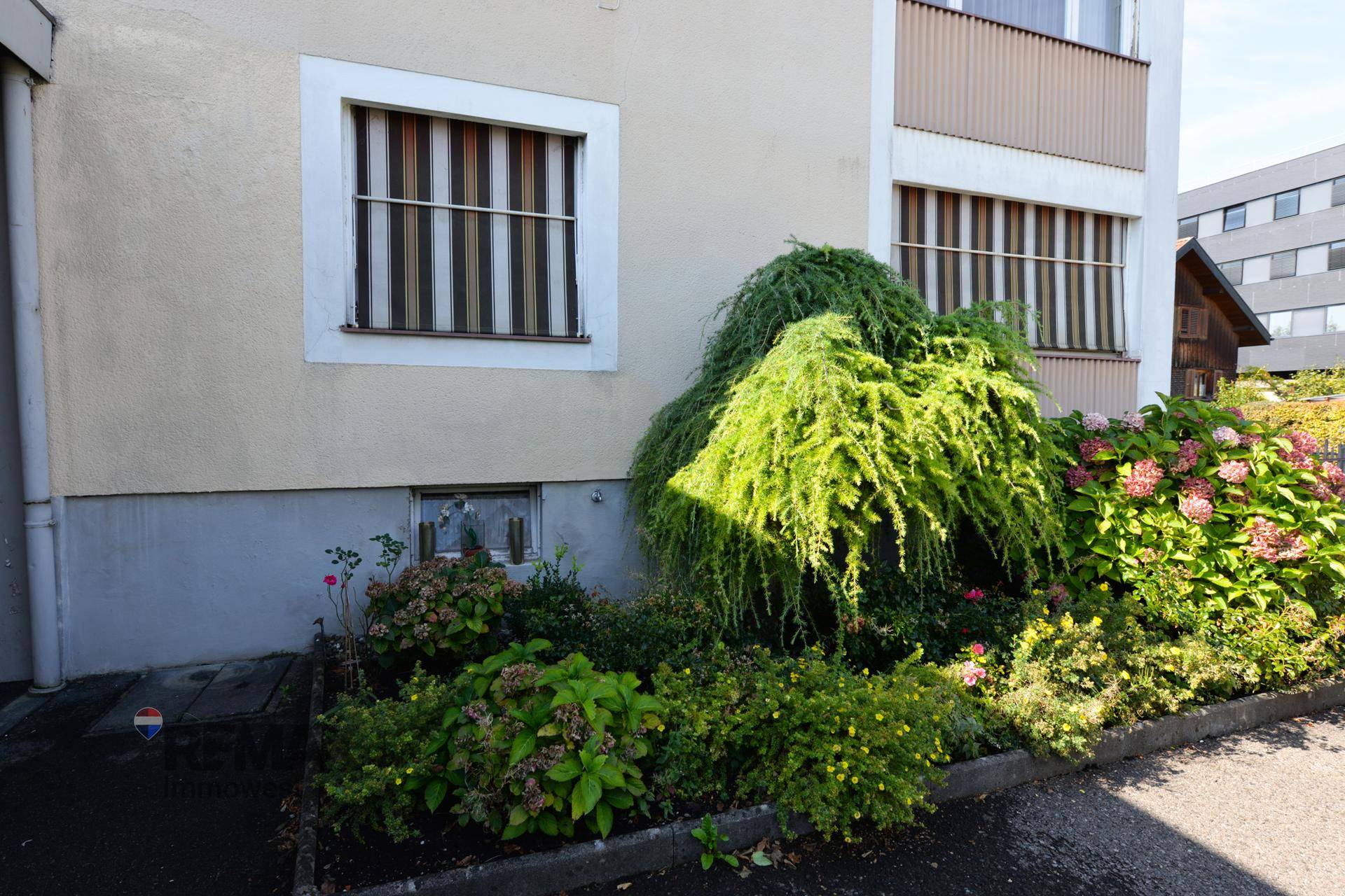 Detailansicht der Hausfassade mit Fenstern und gepflegten Sträuchern und Blumenbeeten.