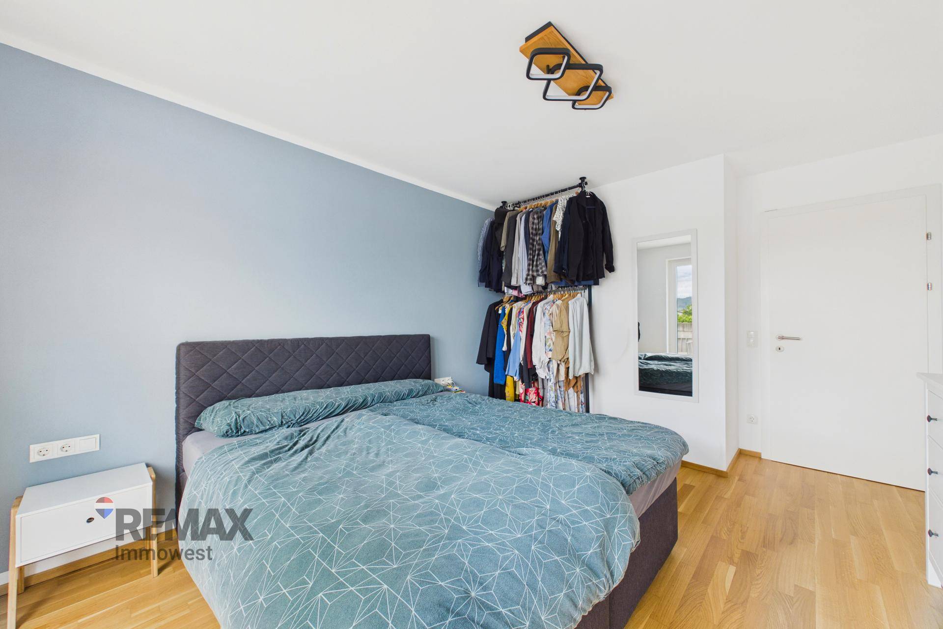 Schlafzimmer mit Doppelbett, offener Kleiderstange und hellem Holzparkettboden.