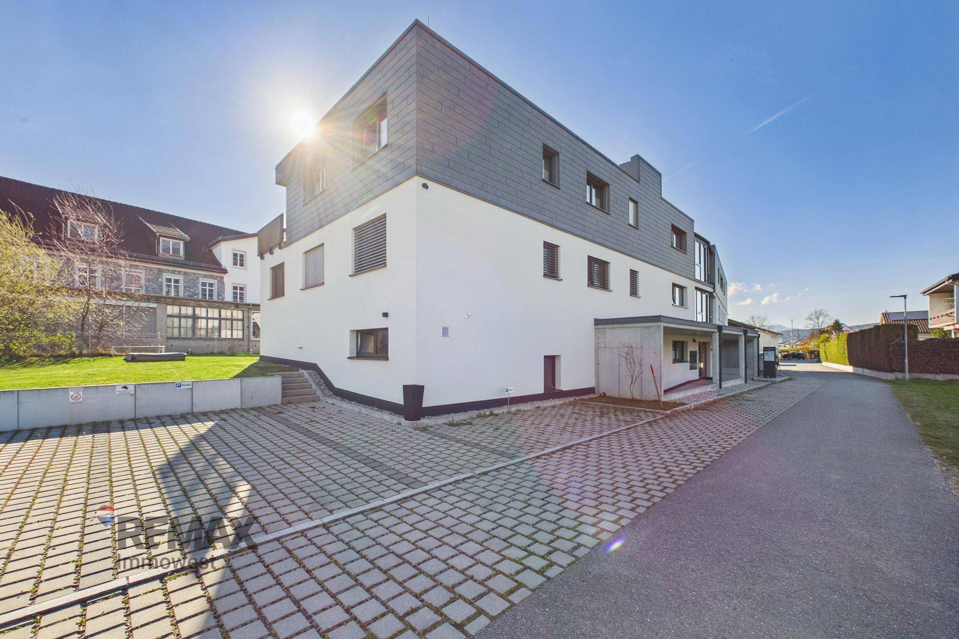 Modernes Gebäude mit Flachdach und gepflastertem Vorplatz unter strahlendem Sonnenschein.