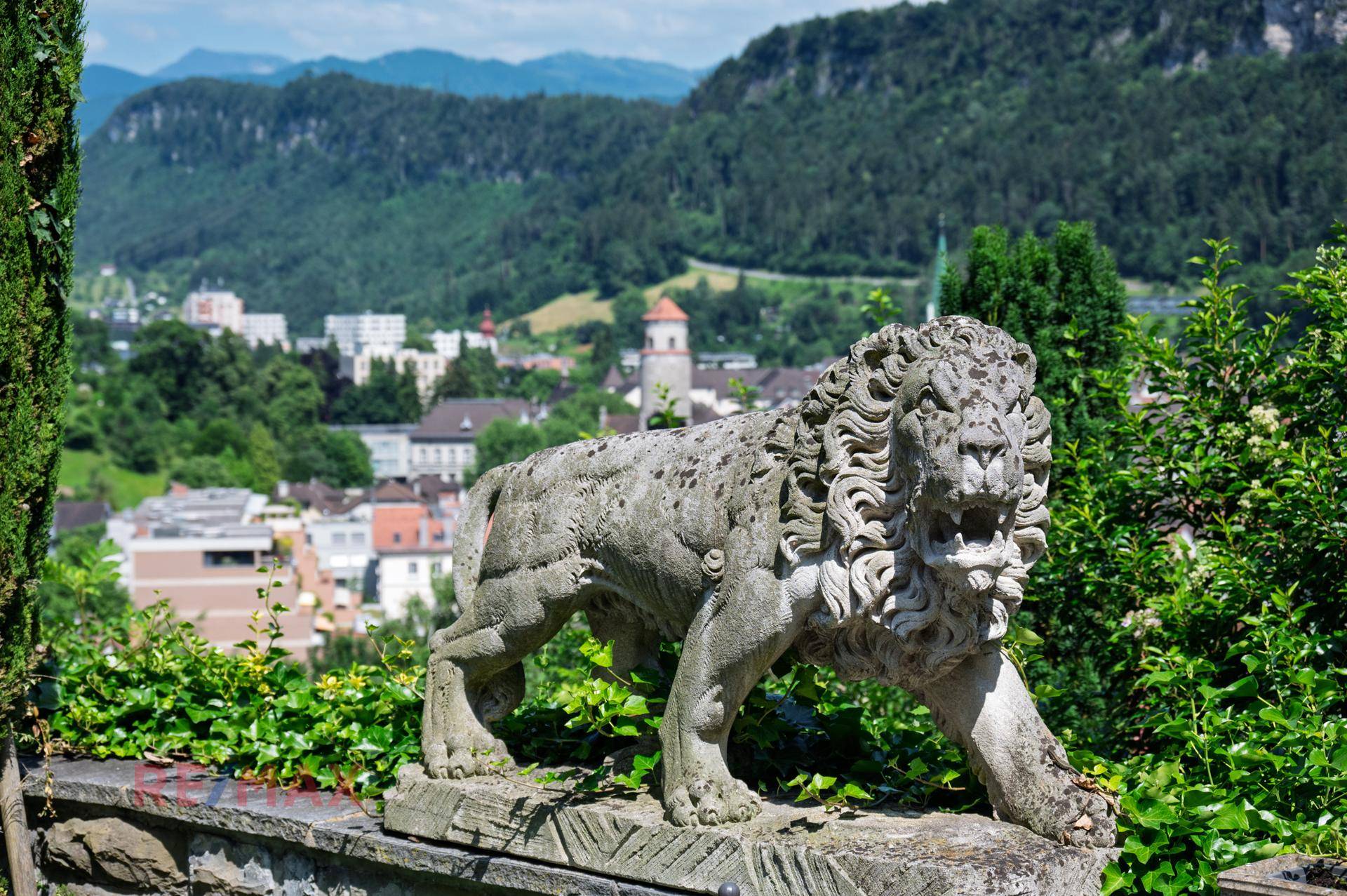 Löwenstatue im Garten mit Panoramablick auf die Stadt und die bewaldeten Berge.