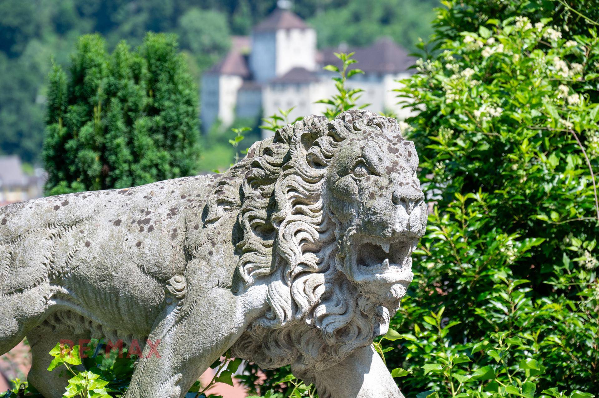 Detailaufnahme einer Löwenstatue im Garten mit historischem Schloss im Hintergrund.