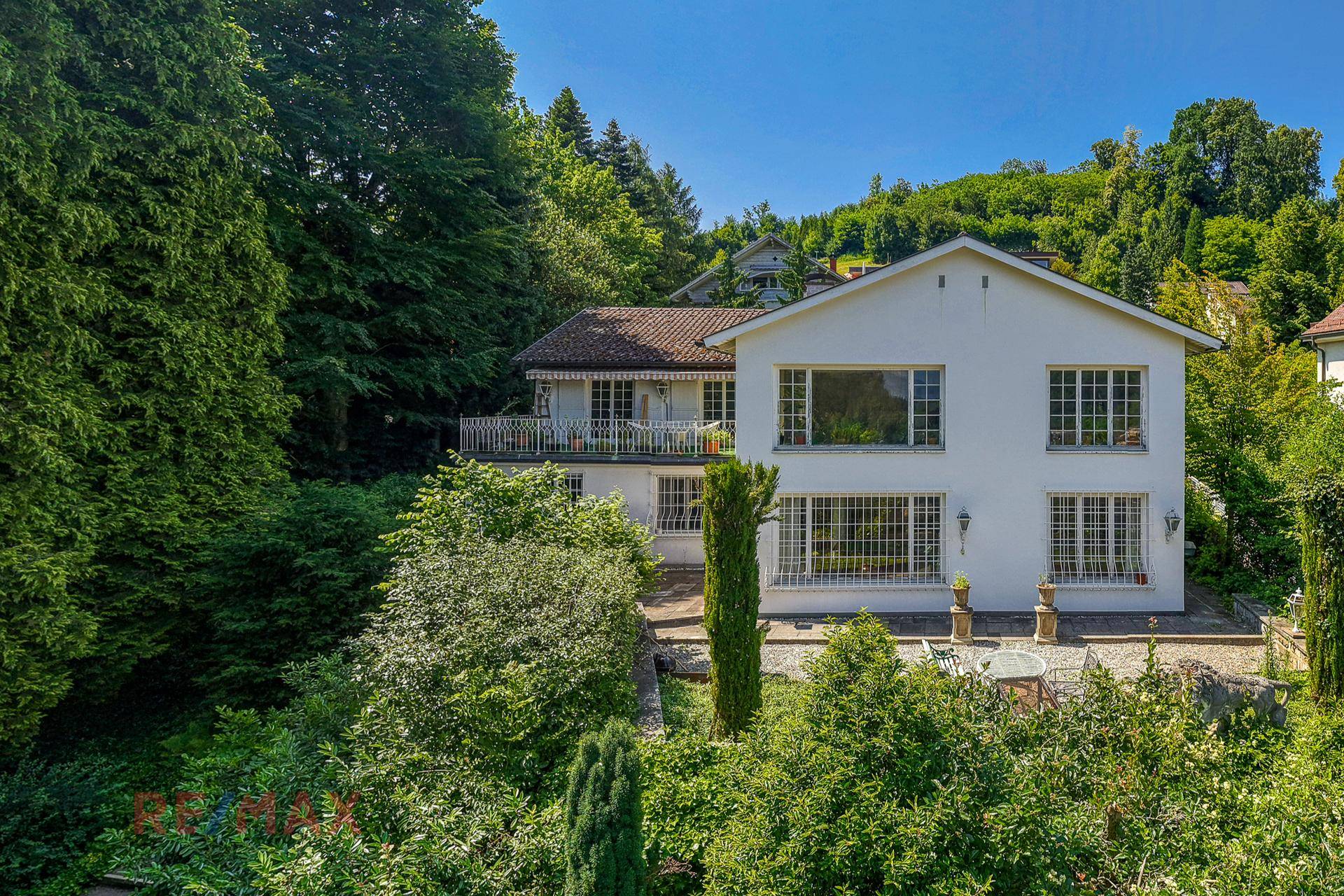 Weiße Villa umgeben von üppigem Grün und hohen Bäumen unter strahlend blauem Himmel.
