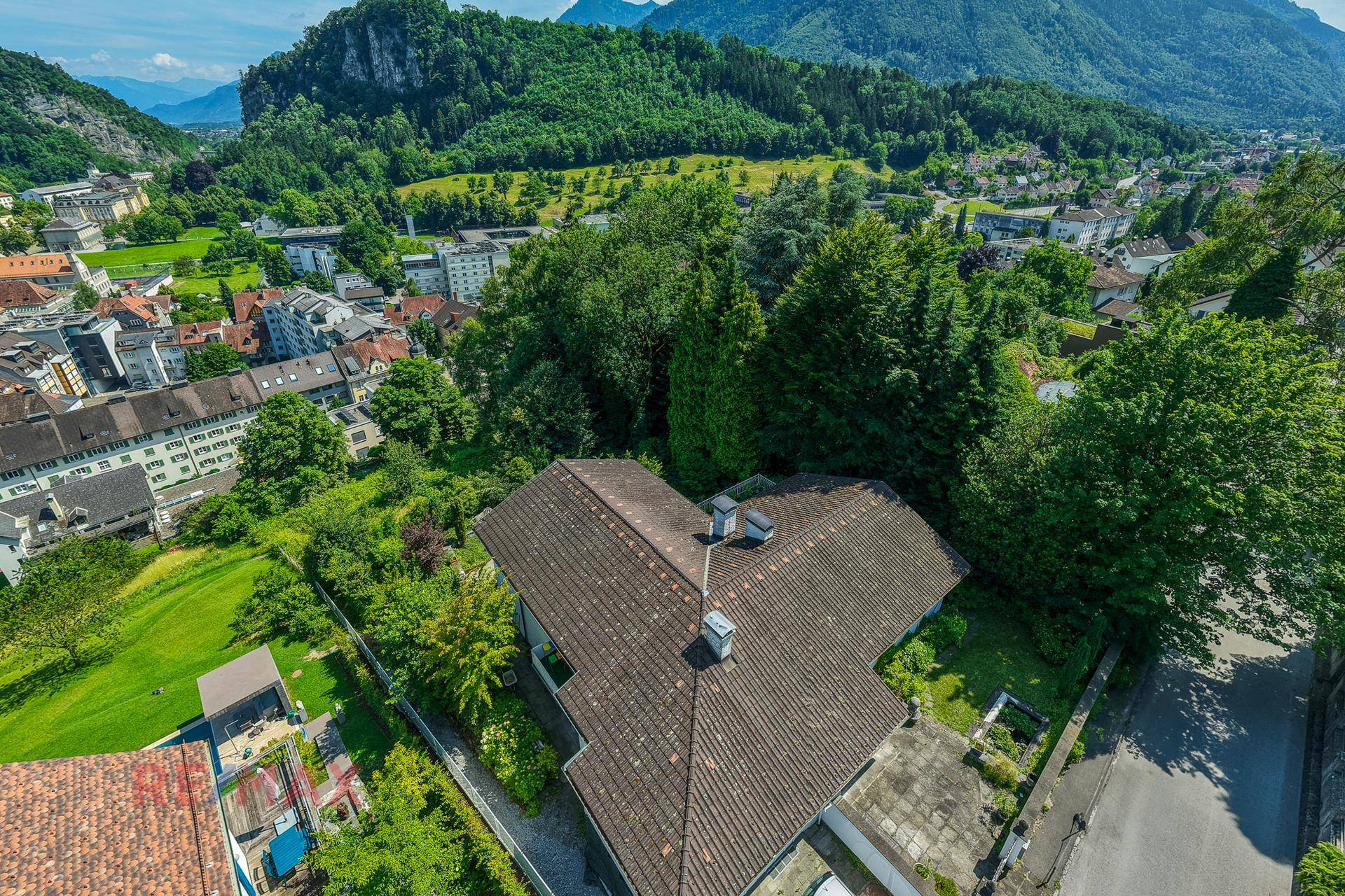 Luftaufnahme eines Hauses mit Ziegeldach, umgeben von Natur, mit Blick auf eine Stadt und Berge.
