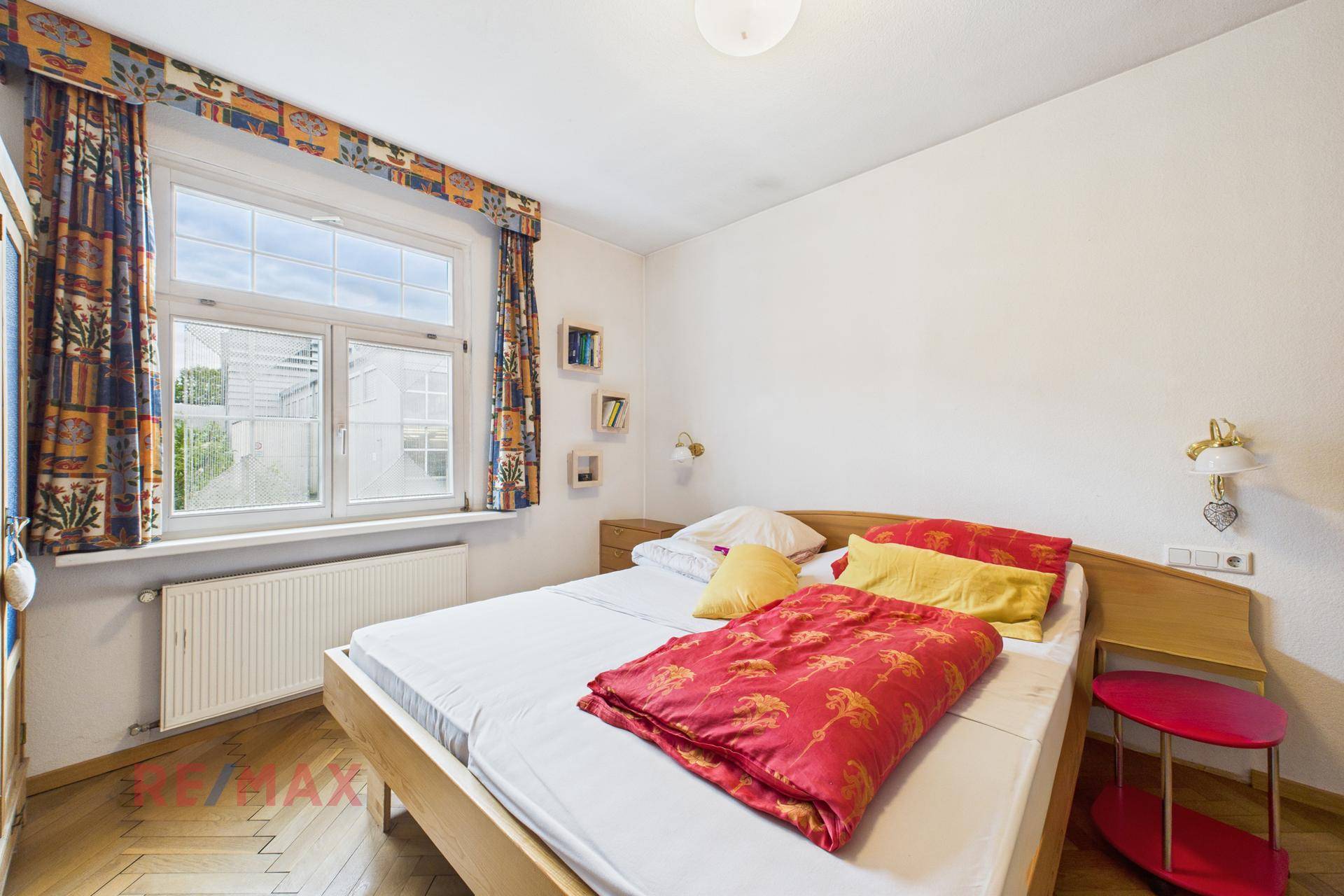 Schlafzimmer mit Doppelbett, Parkettboden und bunten Vorhängen am Fenster.