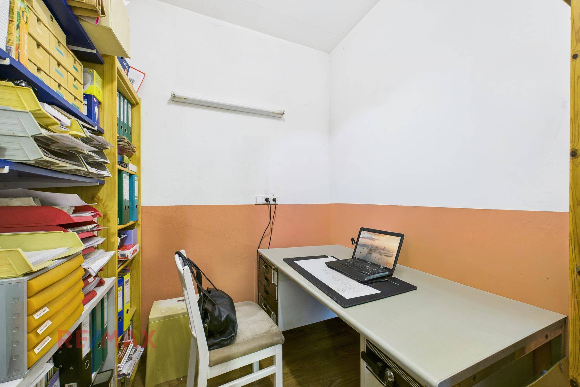 Kleines Büro mit Schreibtisch, Laptop und Regalen voller Aktenordner.
