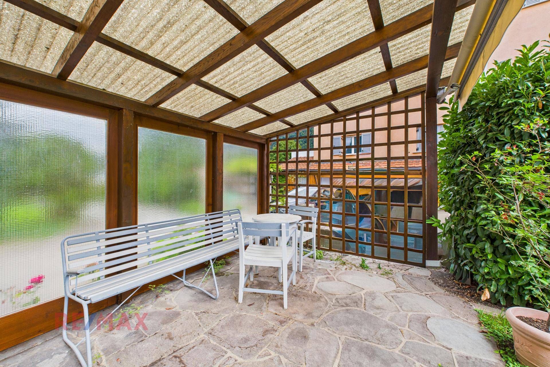 Überdachte Terrasse mit Steinboden und Sitzgelegenheiten, umgeben von grüner Bepflanzung.