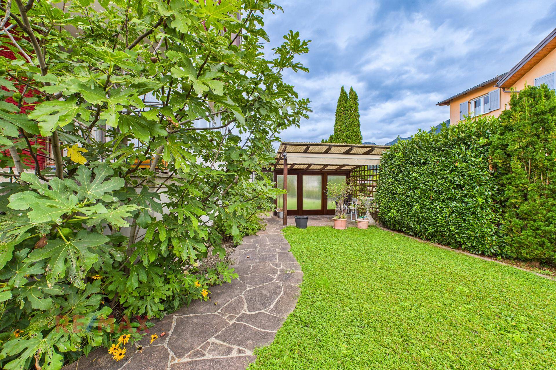 Grüner Garten mit Steinweg, Rasenfläche und überdachtem Sitzbereich im Hintergrund.