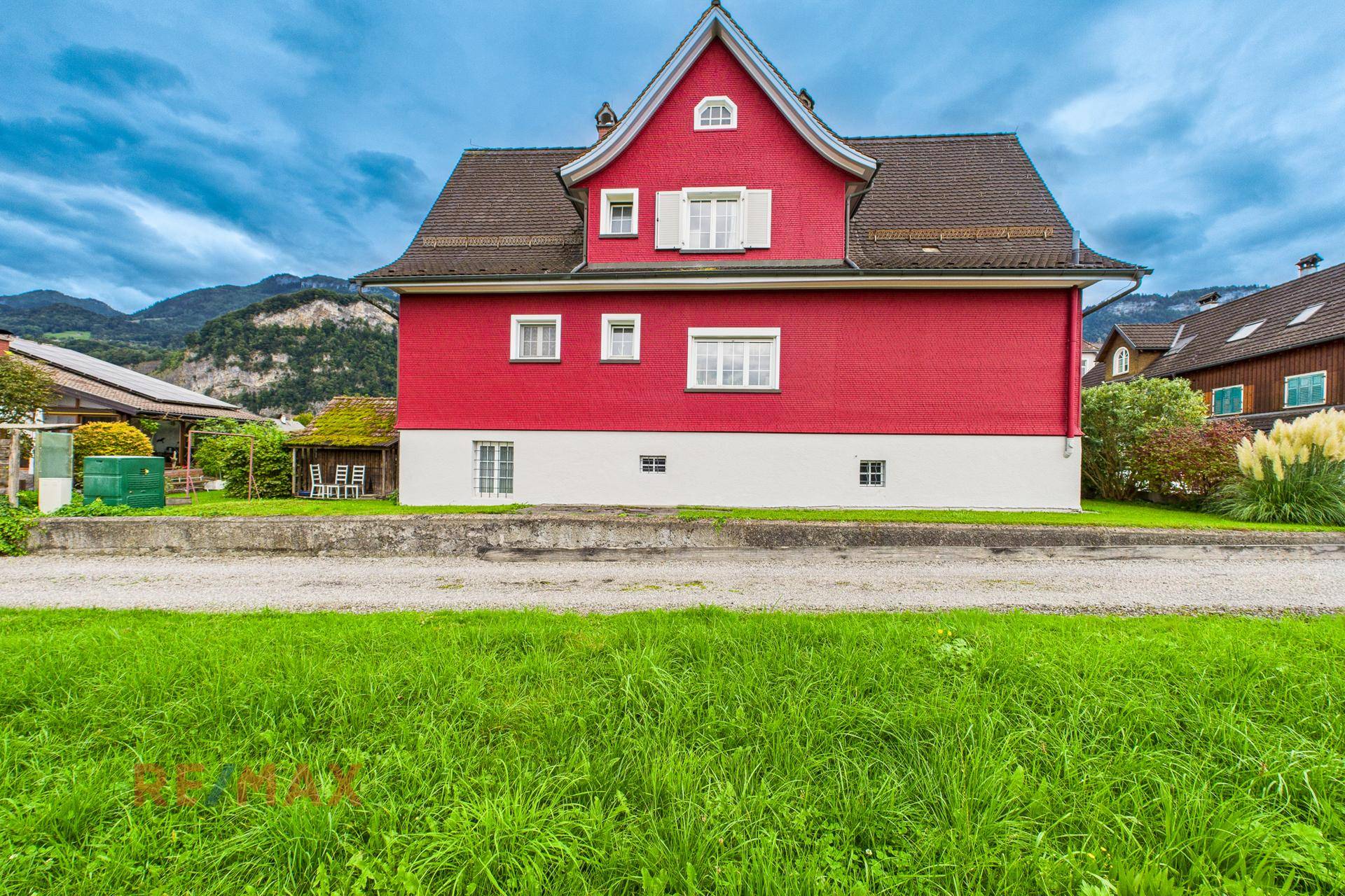 Rotes Wohnhaus mit weißem Sockel und Satteldach, umgeben von grüner Landschaft.
