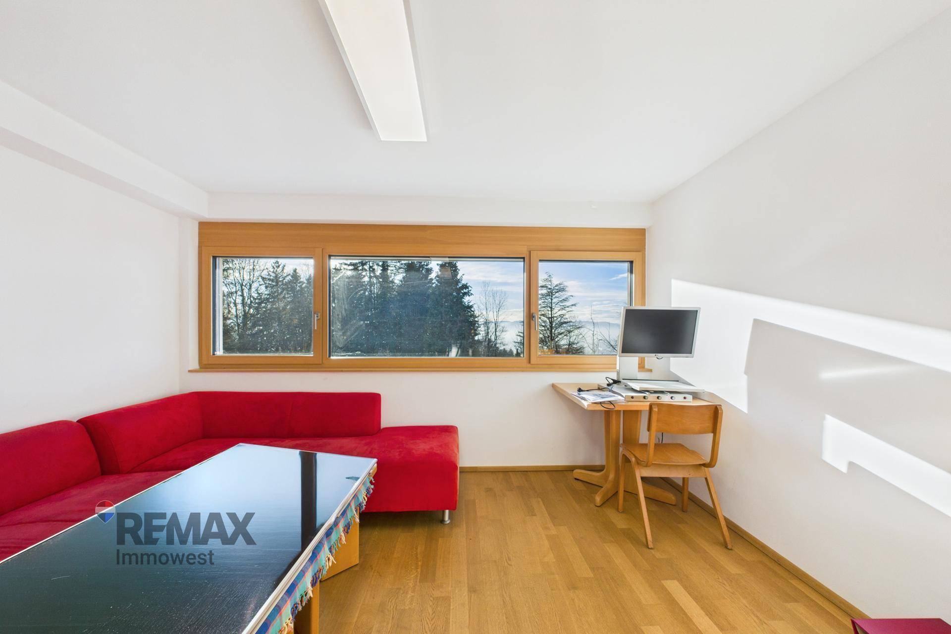 Heller Wohnraum mit Holzboden, rotem Sofa und einem Arbeitsbereich am Fenster mit Waldblick.