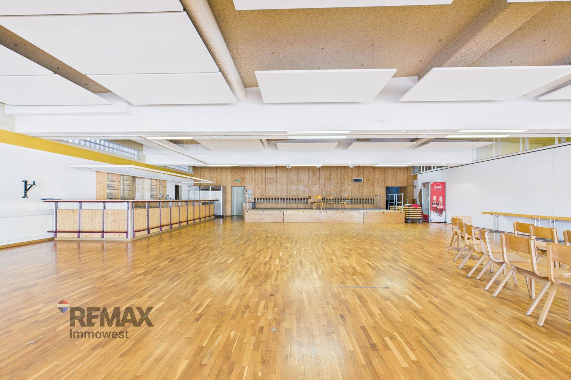 Großer Veranstaltungssaal mit Holzboden, Bühne und einer langen Bar, ideal für Aufführungen und Feste.