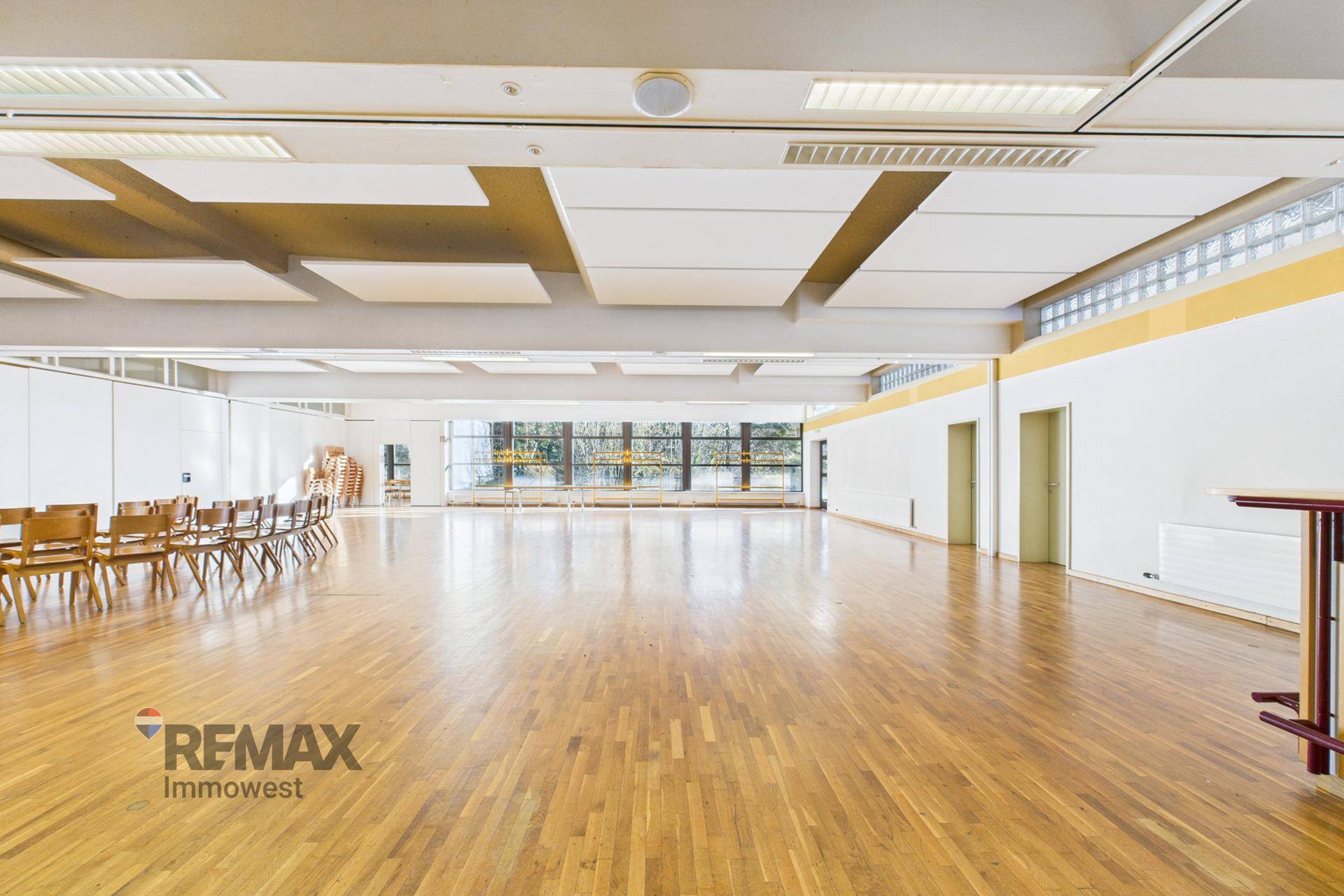 Großer, heller Saal mit Holzboden und hohen Decken, ausgestattet mit Stühlen und großen Fenstern.