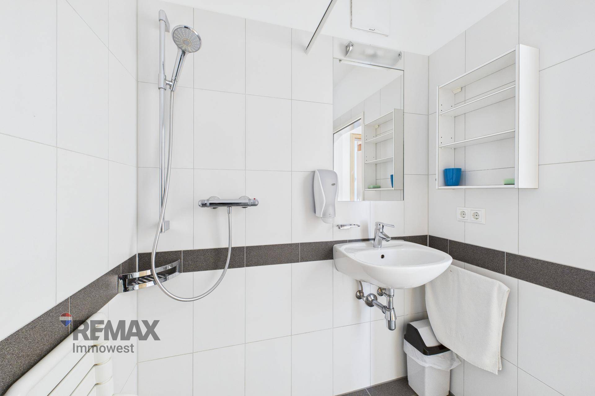 Modernes Badezimmer mit Dusche, Waschbecken und weißen Fliesen, akzentuiert durch eine graue Bordüre.