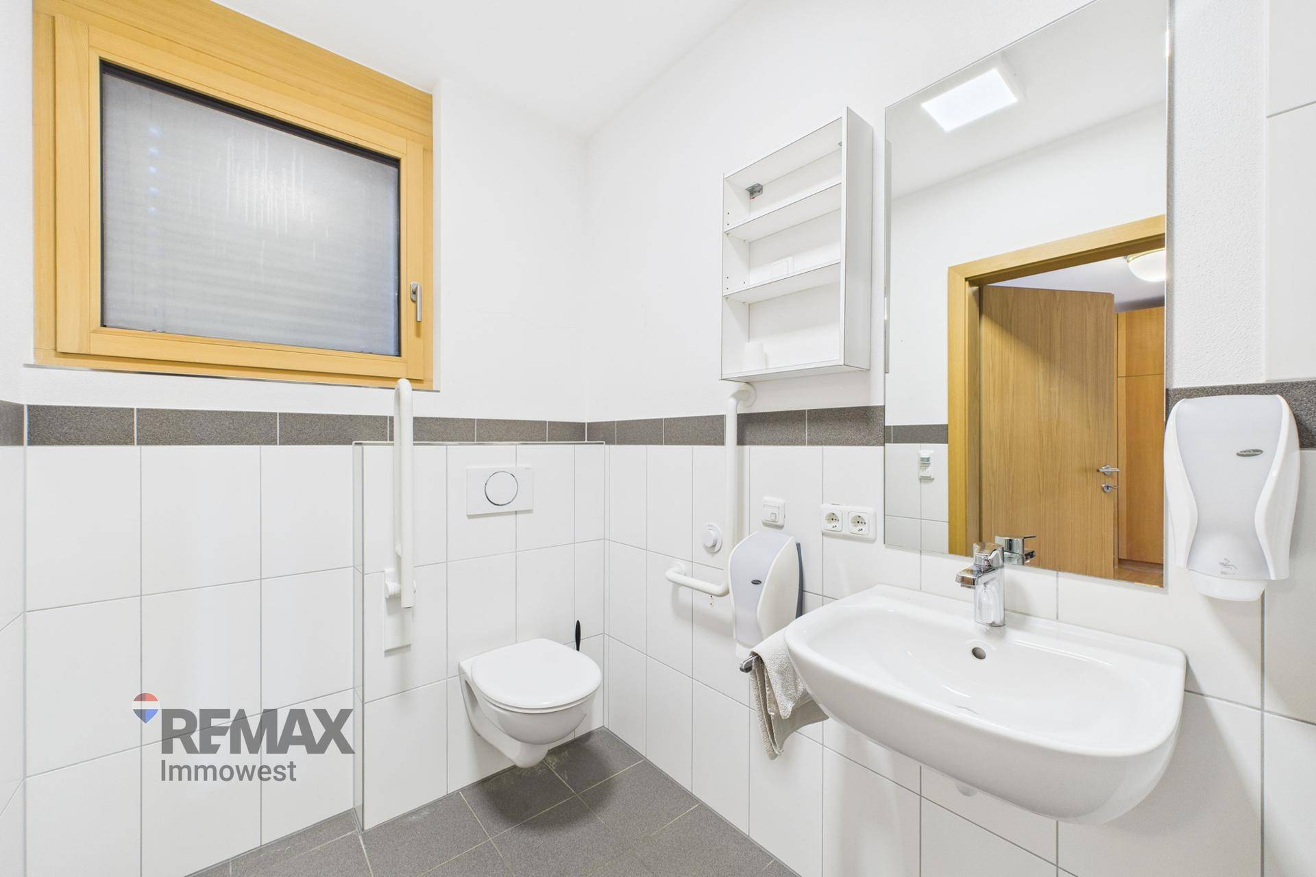 Helles Badezimmer mit WC, Waschbecken und Fenster, ausgestattet mit weißen Fliesen und grauer Bordüre.