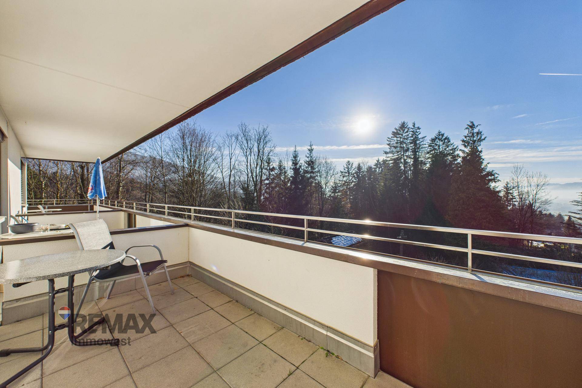 Breiter Balkon mit Fliesenboden und Sitzgelegenheiten, bietet einen sonnigen Ausblick auf die Baumlandschaft.