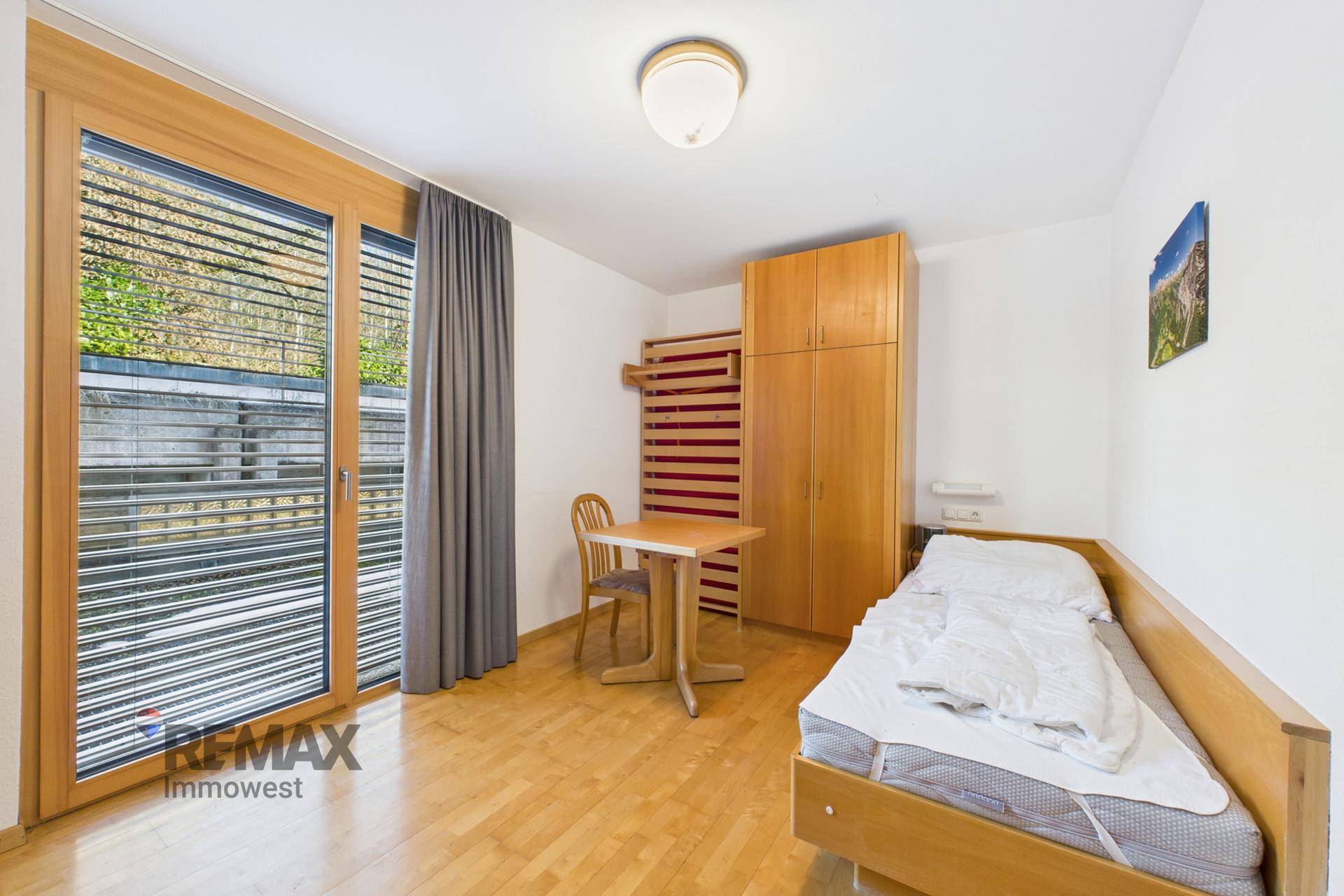 Einzelzimmer mit Bett, Holzschrank und großem Fenster mit Zugang zu einem privaten Balkon.