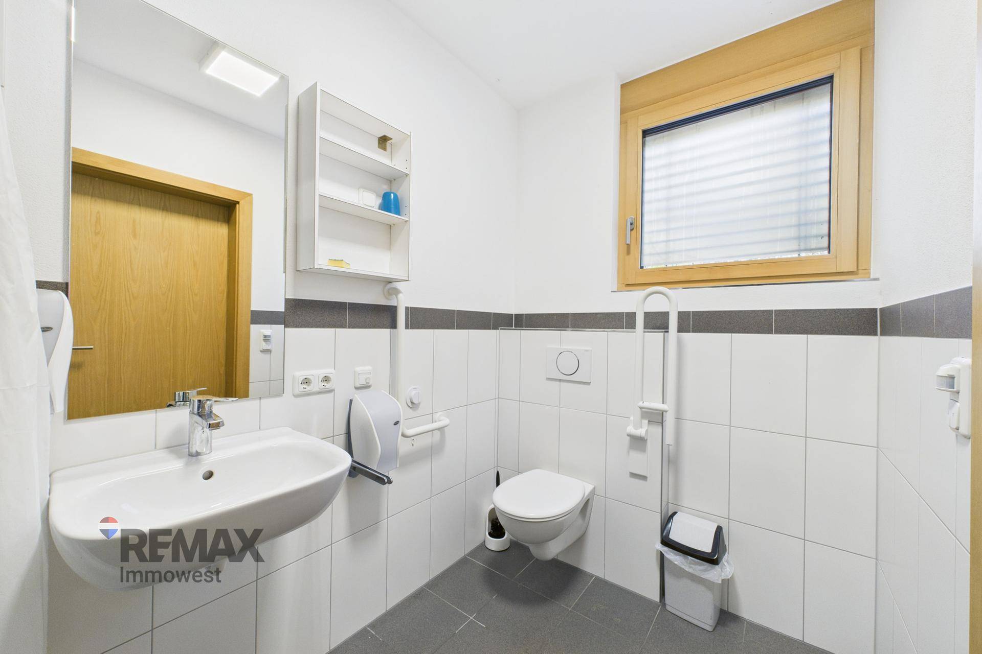 Funktionales Badezimmer mit weißen Fliesen, Waschbecken, Toilette und Fenster, ausgestattet mit Haltegriffen.