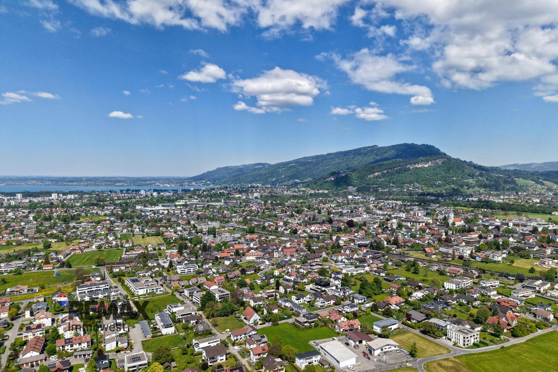 Weitläufige Luftaufnahme einer Stadt am Seeufer mit Bergen im Hintergrund unter blauem Himmel.