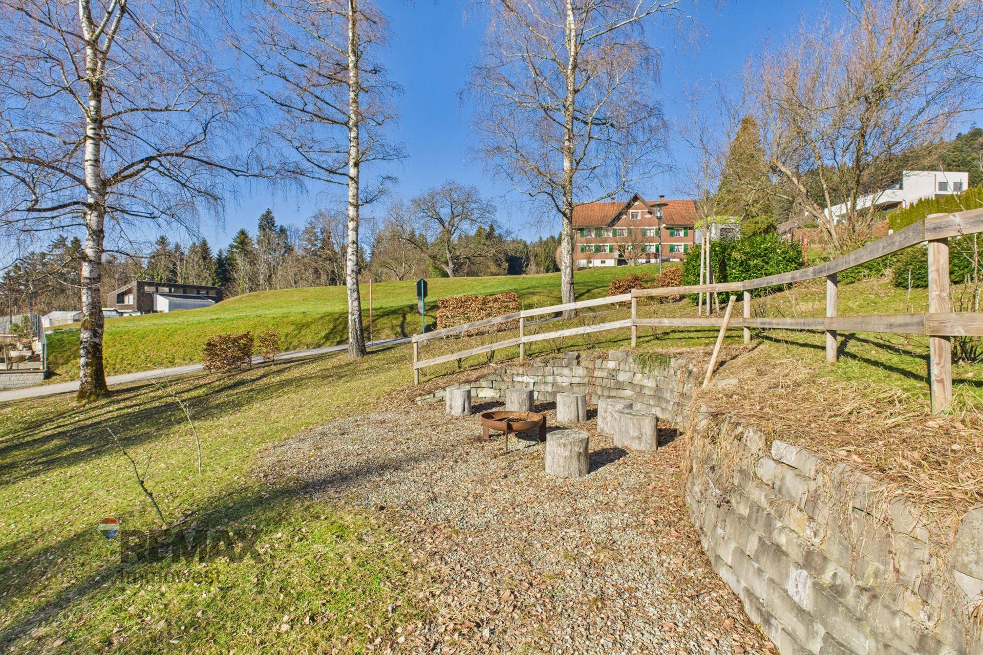 Gestalteter Gartenbereich mit Steinmauer, Holzgeländer und Sitzgelegenheiten für ein Lagerfeuer.