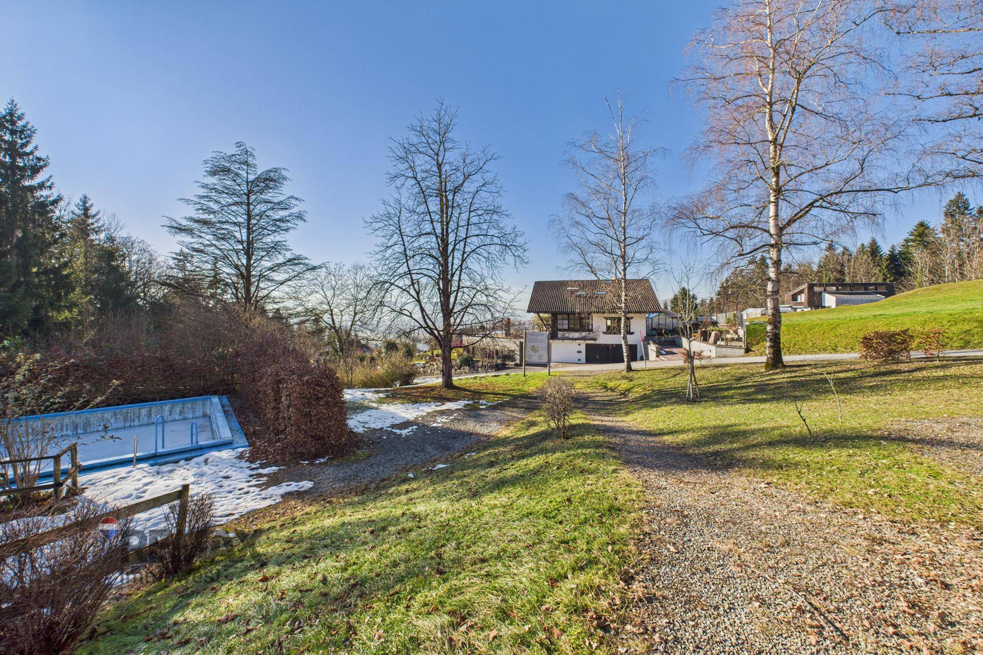 Gartenlandschaft mit einem leeren Pool und einem Haus im Hintergrund unter blauem Himmel.