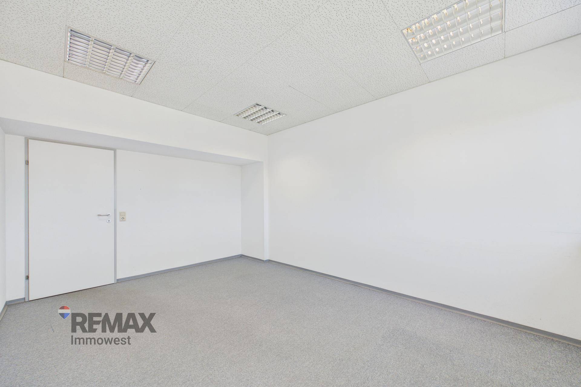 Leeres Büro mit grauem Teppichboden und weißer Tür, ausgestattet mit effizienter Deckenbeleuchtung.