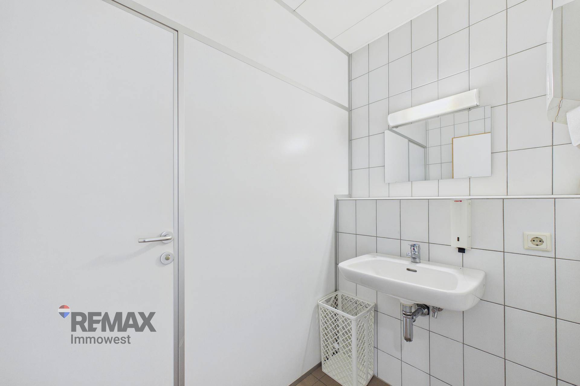 Einfache Toilette mit weißen Fliesen, Waschbecken und Spiegel, funktional und sauber gehalten.