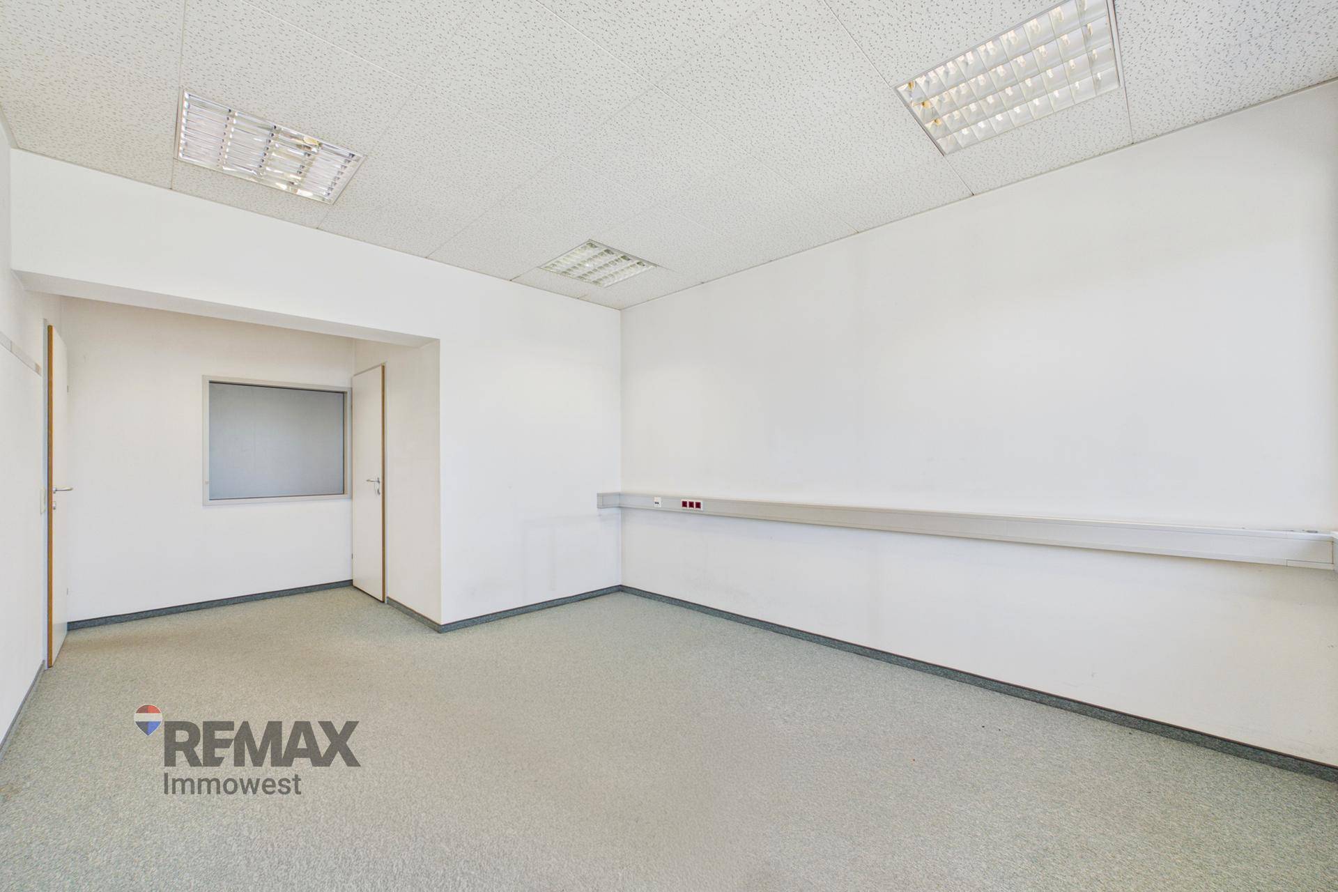 Leeres Büro mit grauem Teppichboden, weißer Tür und einer praktischen Wandablage für Kabel.