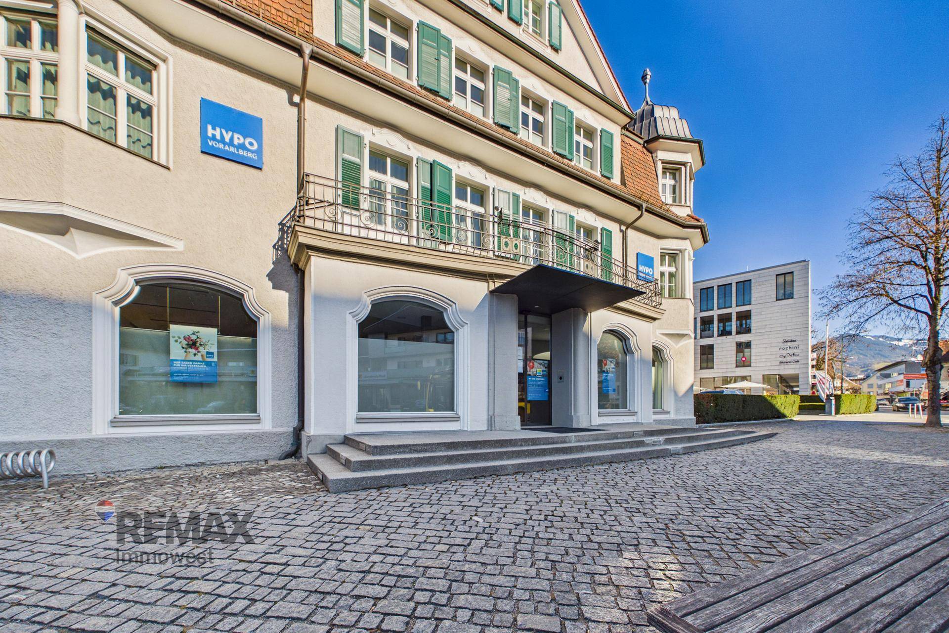 Eingangsbereich des Gebäudes mit Treppe, Balkon und Schaufenstern, umgeben von Kopfsteinpflaster.