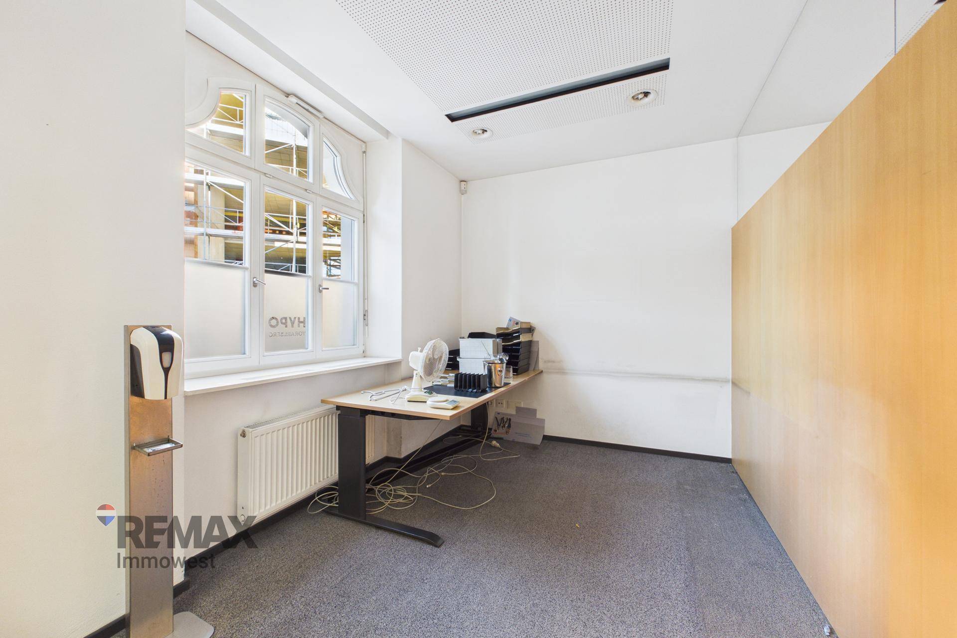 Kleiner Büroraum mit einem Schreibtisch, Heizung unter dem Fenster und einem Spender an der Wand.