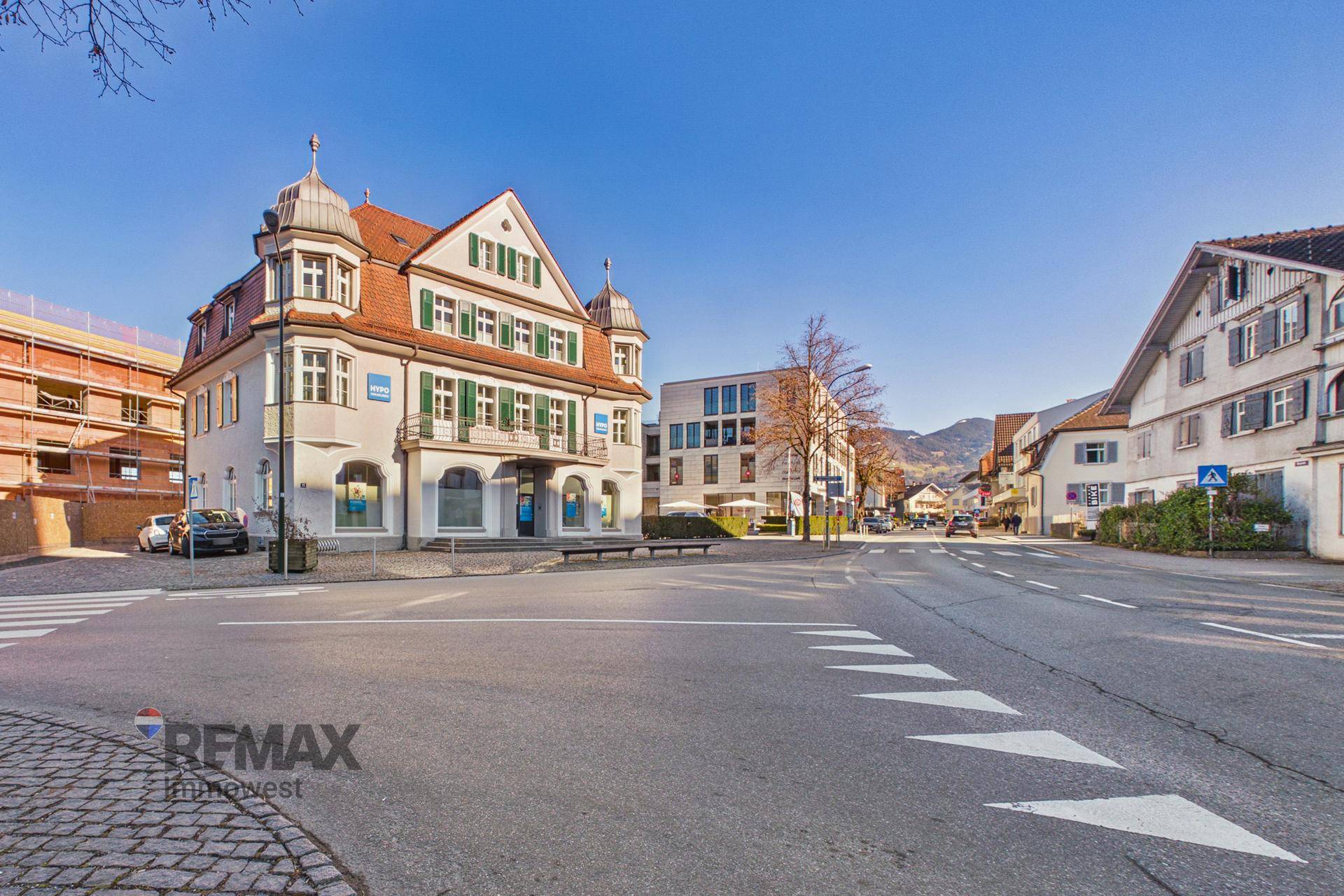 Ansicht des historischen Gebäudes an einer belebten Straße mit umliegender Bebauung und Bergen im Hintergrund.