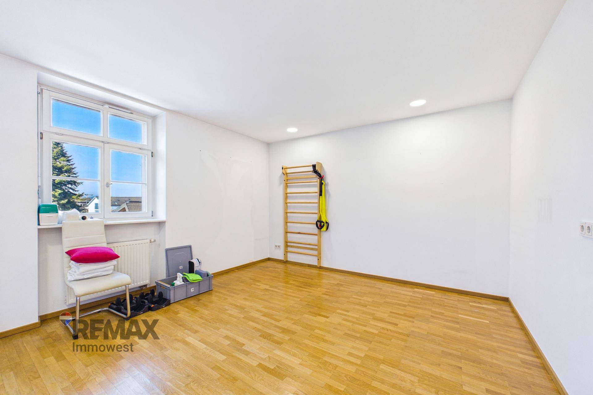 Leerer Therapieraum mit Holzboden, Sprossenwand und großem Fenster, bereit zur Nutzung.