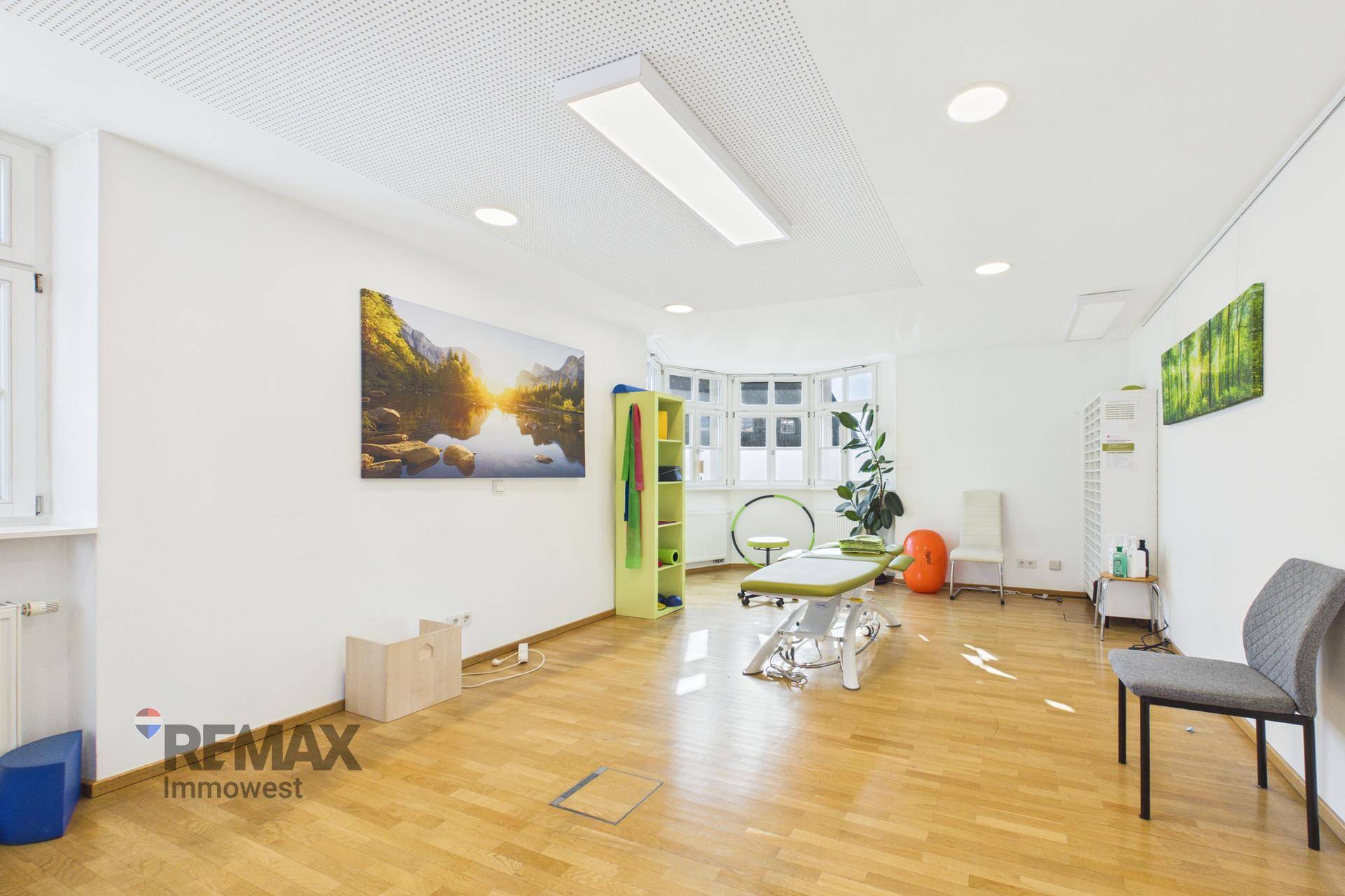 Geräumiger Therapieraum mit Behandlungsliege, Trainingsgeräten und großen Fenstern, hell und einladend.